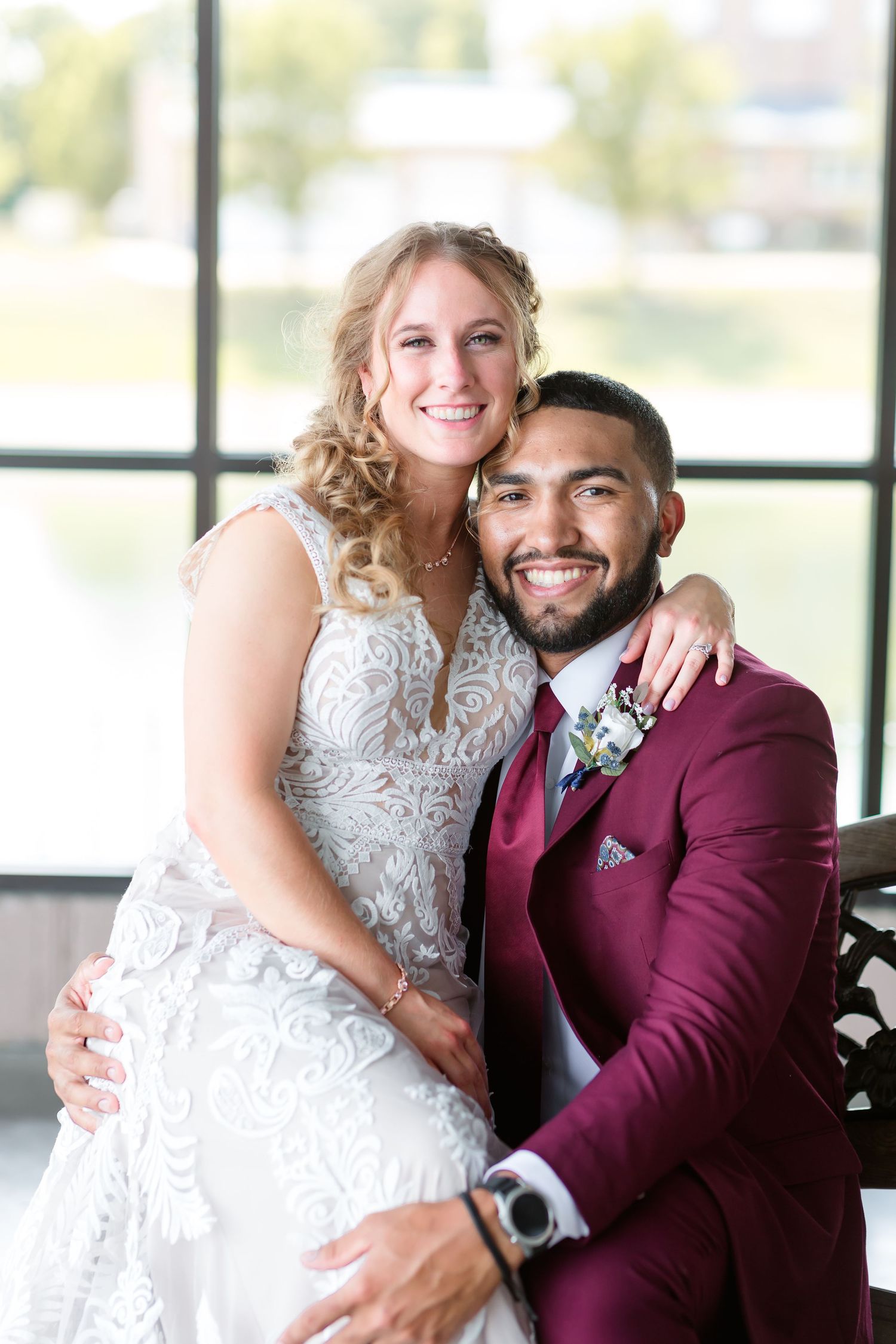 Smiling bride and groom pose together at Lake Lyndsay, groom wearing burgundy suit with boutonniere by large window.