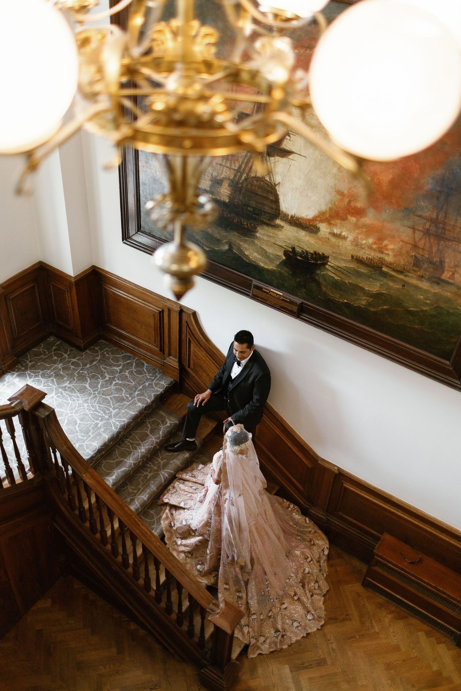 High-angle creative shot of a Muslim couple standing on a grand wooden staircase beneath a classic chandelier at De Vere Wokefield Estate