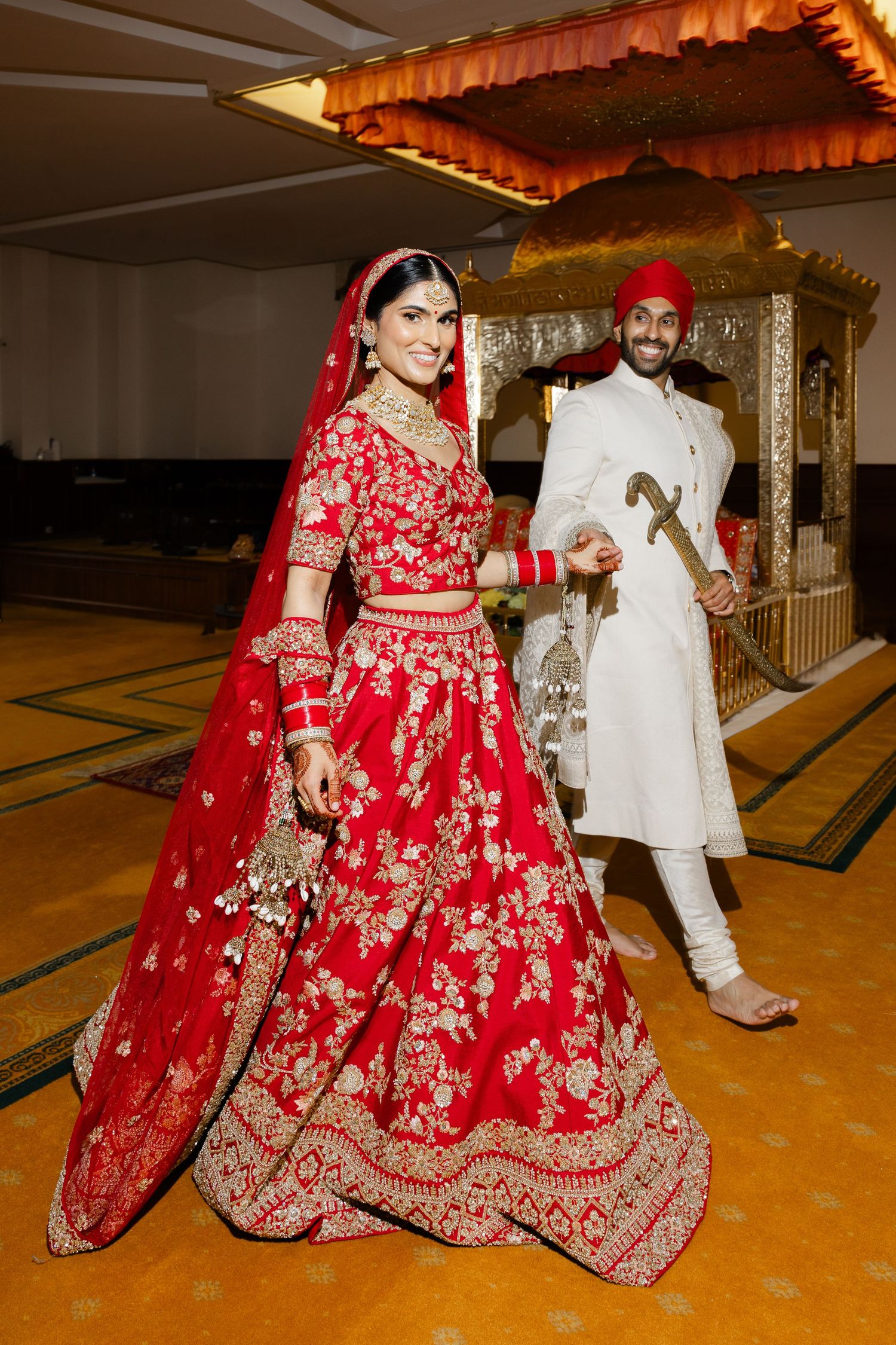 A smiling Sikh bride in a red and gold embroidered lehenga and a groom in a white sherwani walking in Hounslow Gurdwara