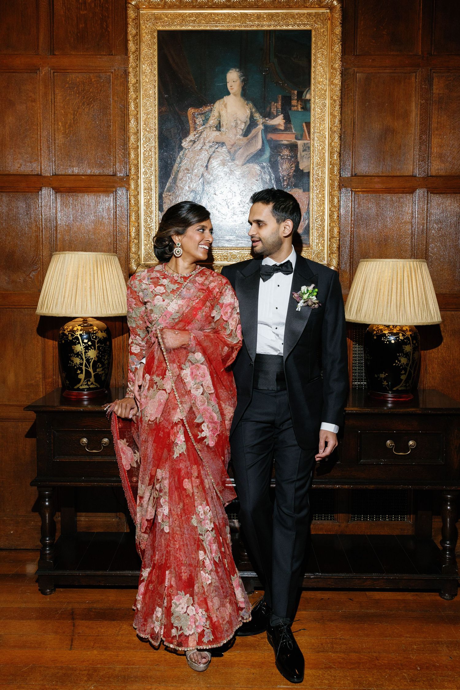 Editorial-stylel indoor portrait of a bride in a red floral sari and a groom in a black tuxedo, standing in North Mymms Park