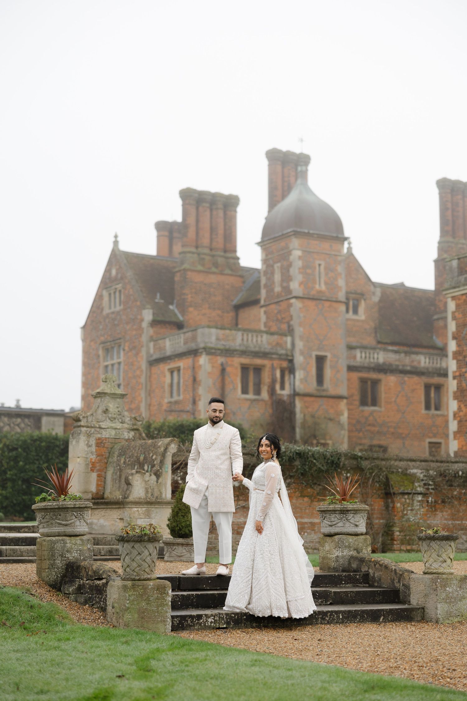 Full-length portrait of a couple in ivory traditional attire standing on stone steps in front of North Mymms Park