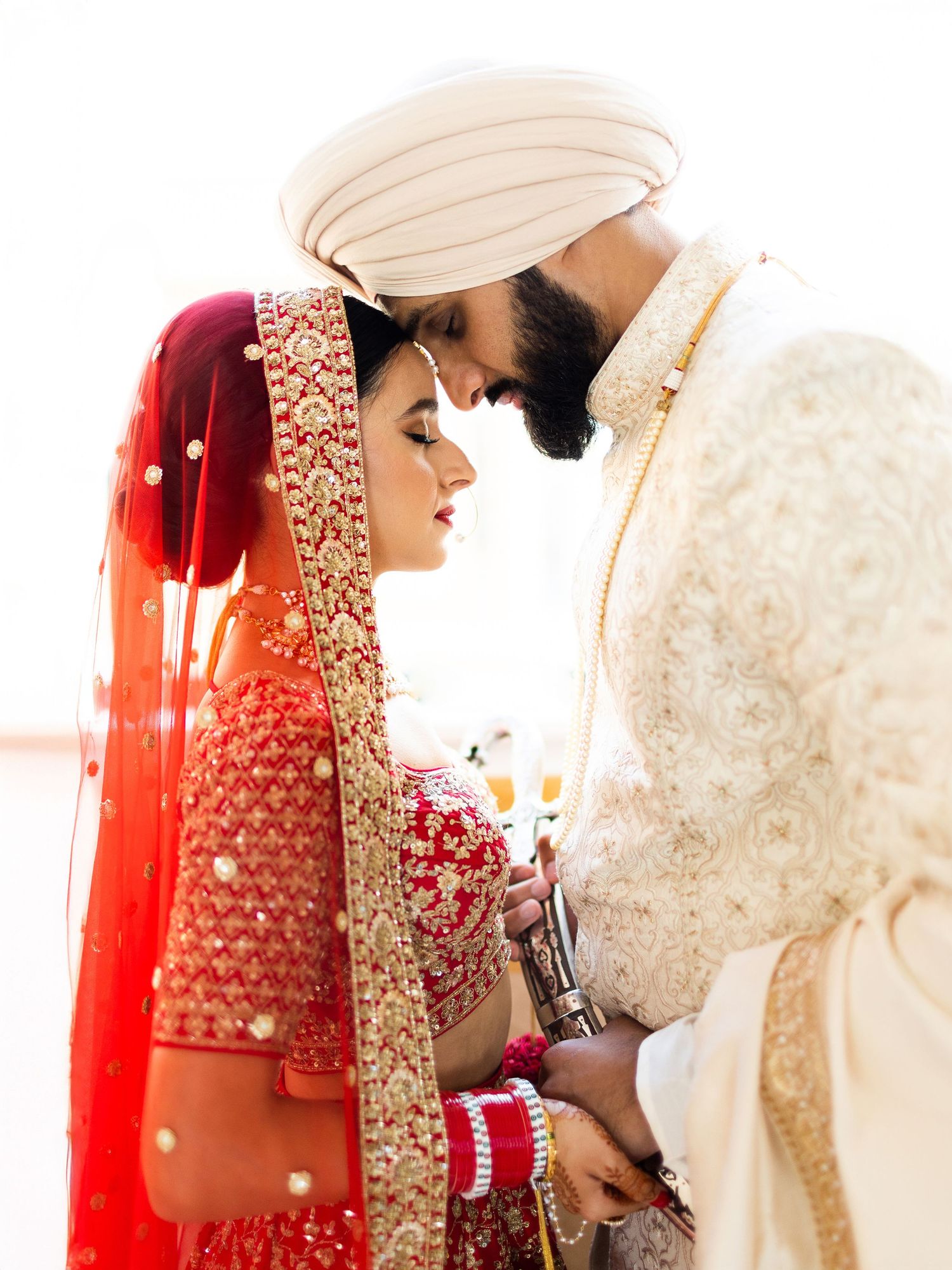 An intimate, high-key side profile portrait of a Sikh bride and groom touching foreheads, showcasing intricate outfit details