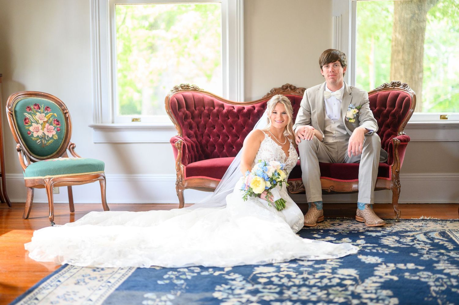 Bride sitting on floor in white gown holding bouquet beside groom on vintage red velvet sofa in elegant room.