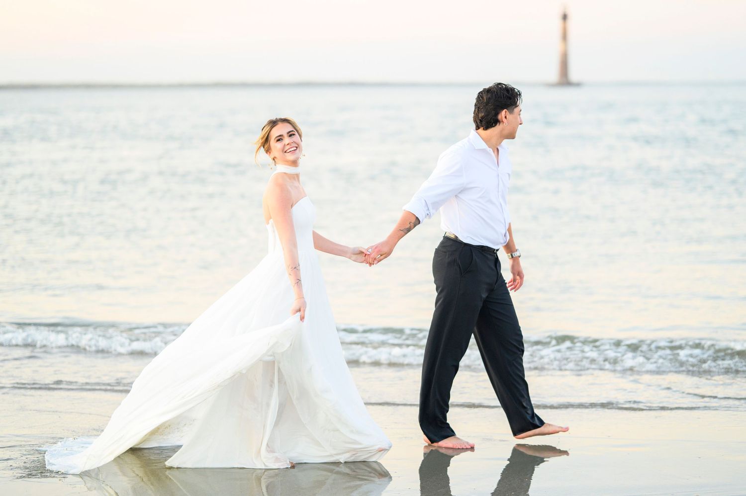 Bride and groom walking hand in hand barefoot on a sandy beach, with ocean waves and a lighthouse in the background.
