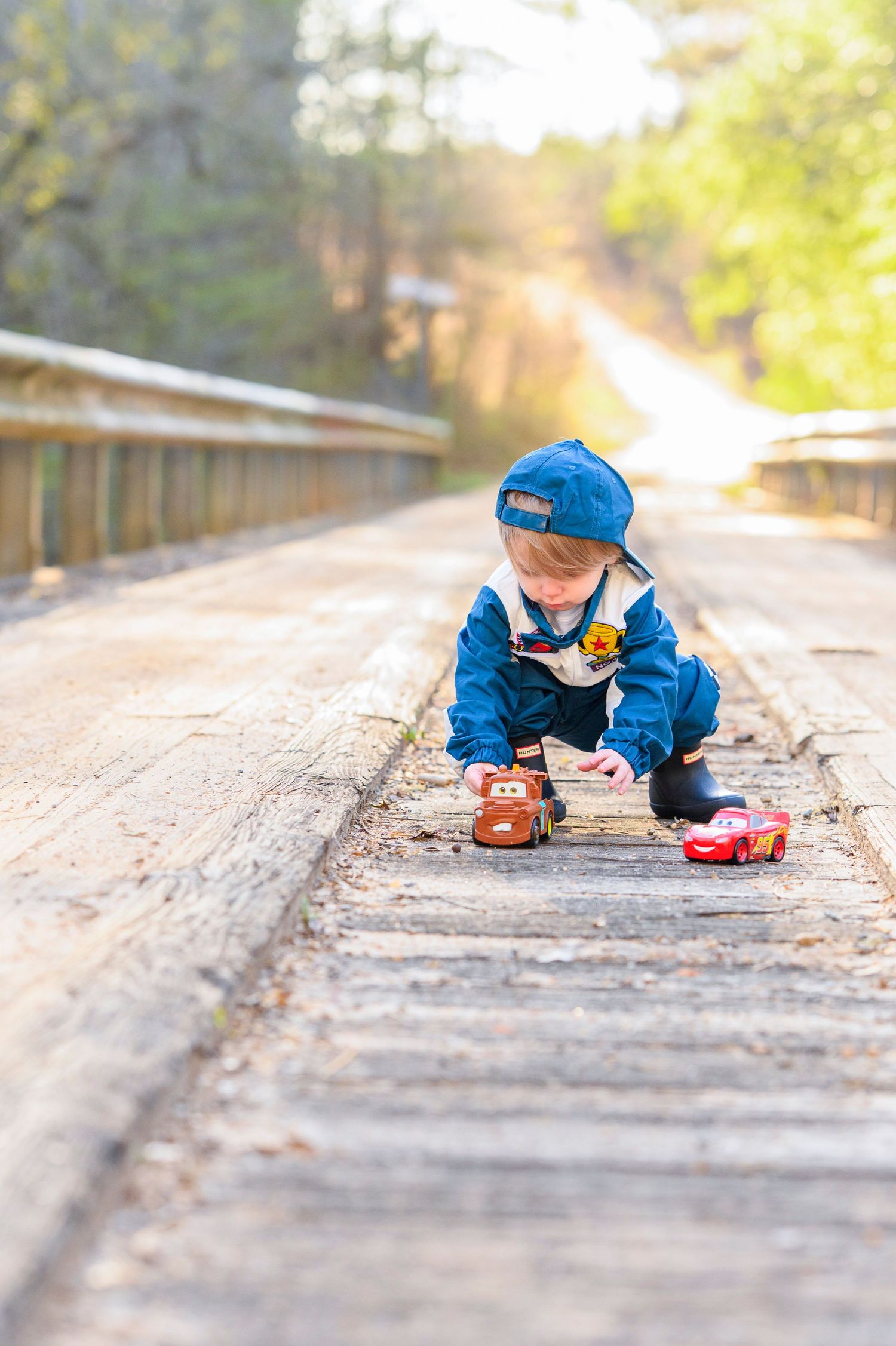 Young boy in blue outfit plays with toy cars on a wooden bridge in sunlight.