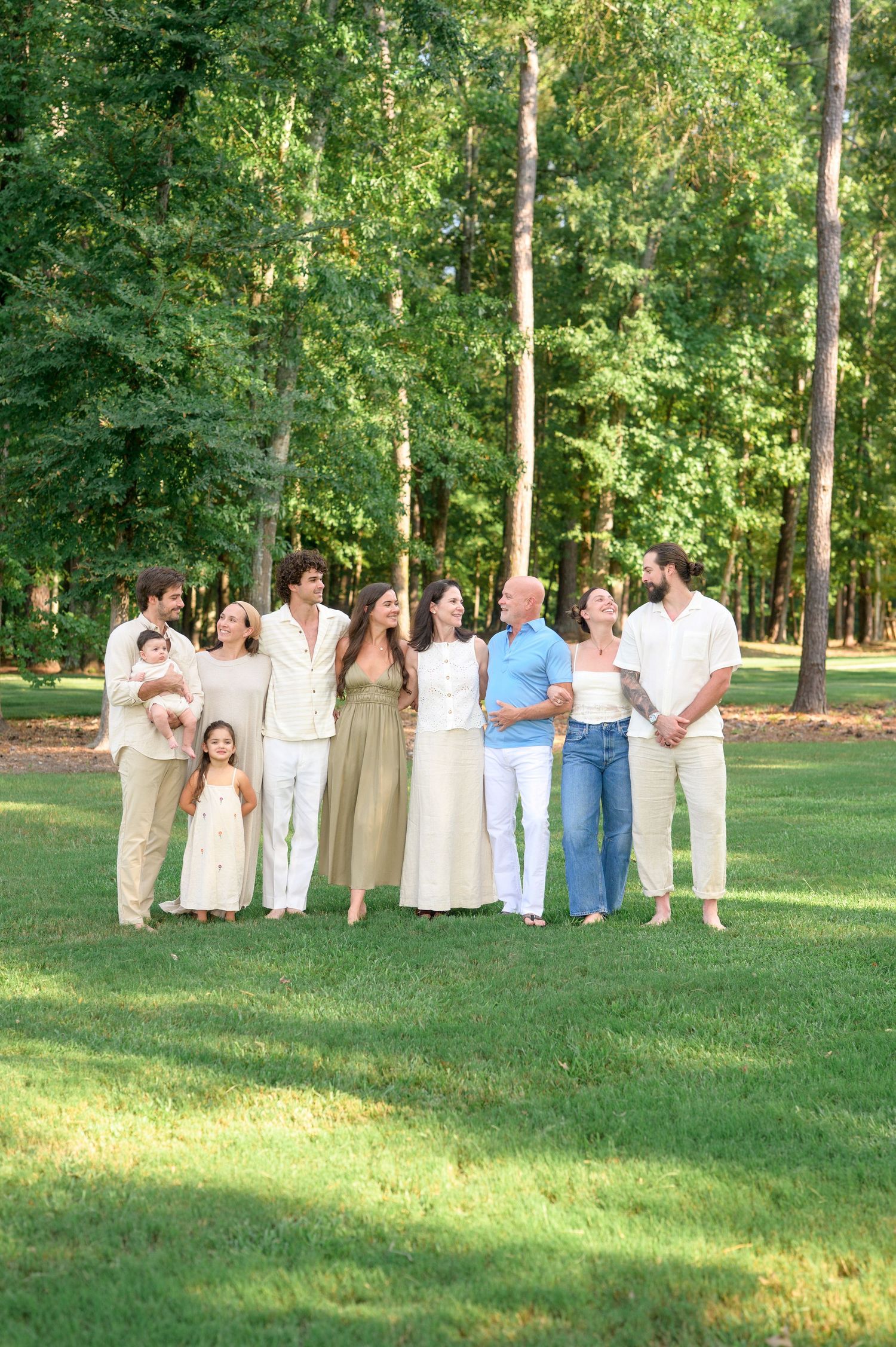 A multigenerational family of nine poses together on a green lawn surrounded by tall trees in a park.