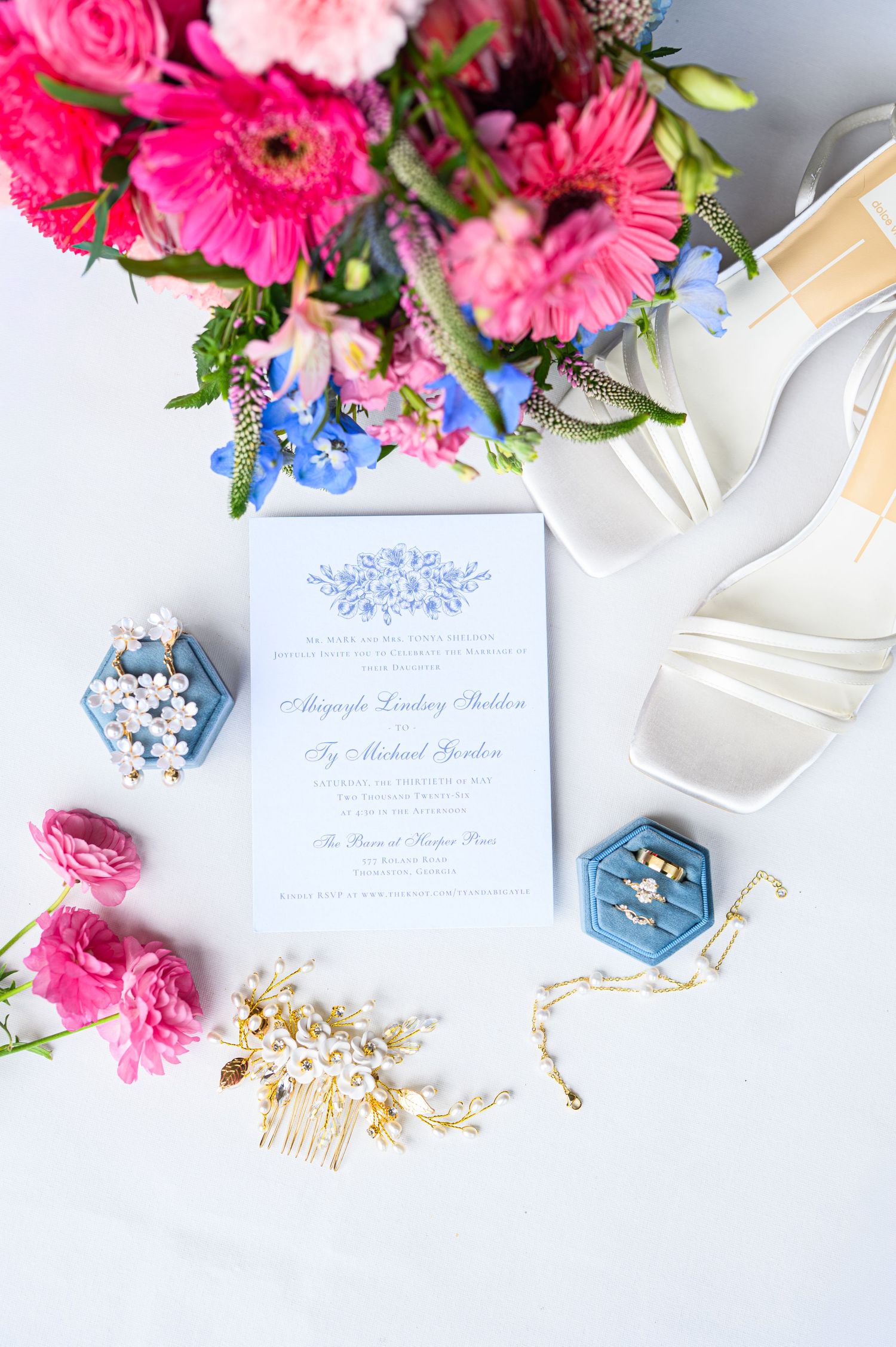 Elegant wedding flat lay with blue invitations, pink floral bouquet, white heels, gold hair comb, and velvet ring boxes.