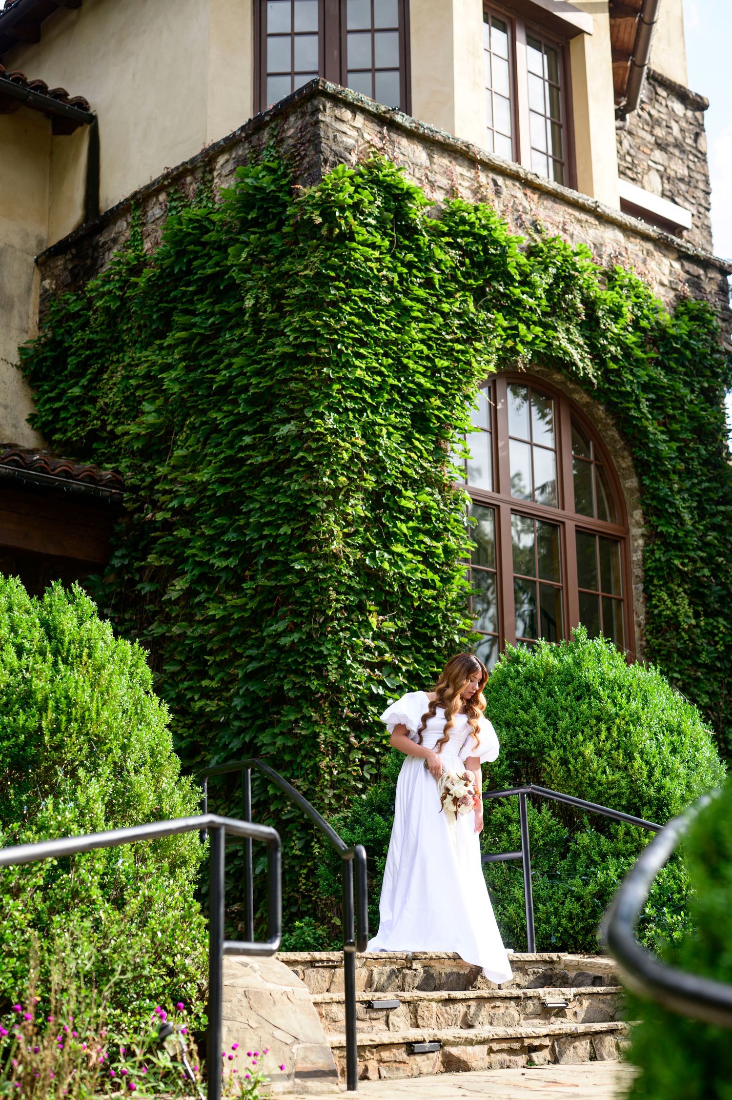 Bride in white gown holding bouquet stands on stone steps before ivy-covered stone building with arched windows.