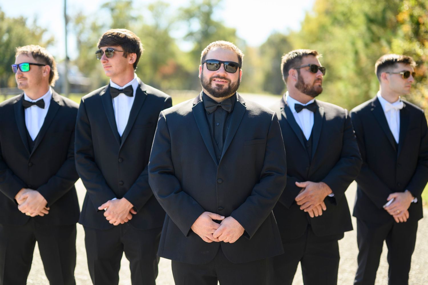 Groom and groomsmen in black tuxedos and bow ties wearing sunglasses outdoors.