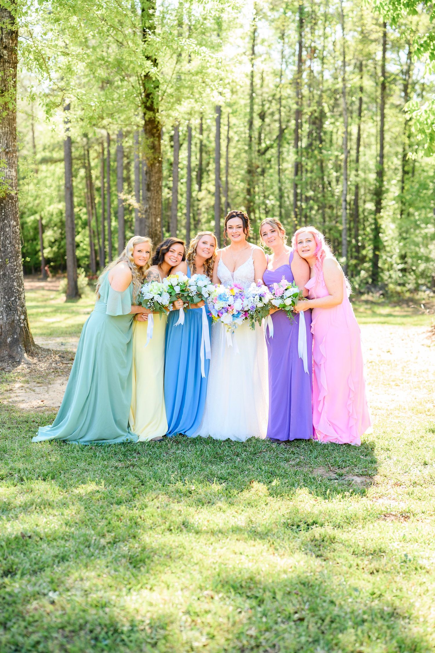 Bride and bridesmaids in rainbow-colored gowns posing in a sunlit forest setting.