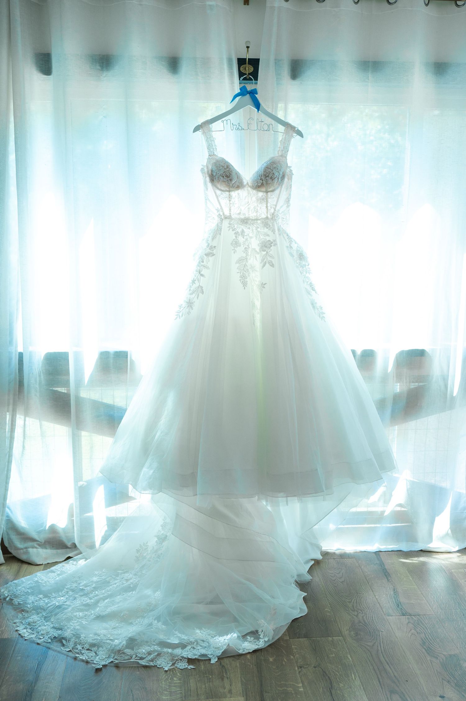 Elegant white bridal ball gown with lace appliques and long train hanging by a window with sheer curtains.