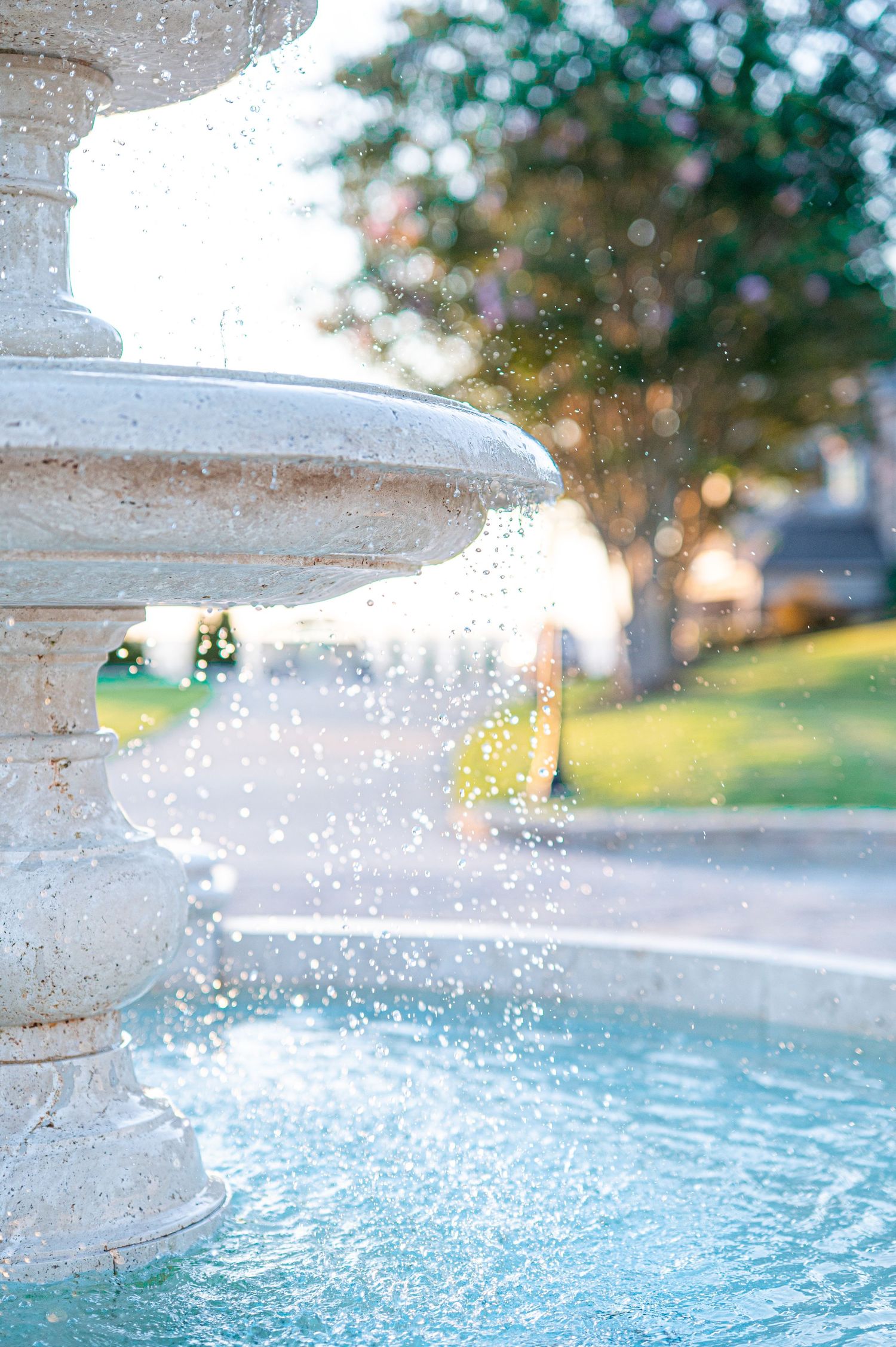Close-up of a white tiered stone fountain with splashing water, surrounded by lush green trees in a sunny park.