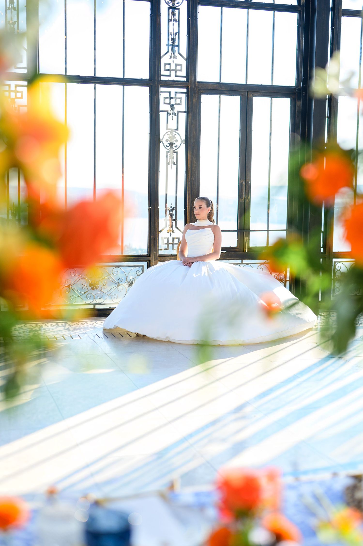 Bride in white ball gown seated by ornate windows, framed by vibrant orange roses in elegant venue.