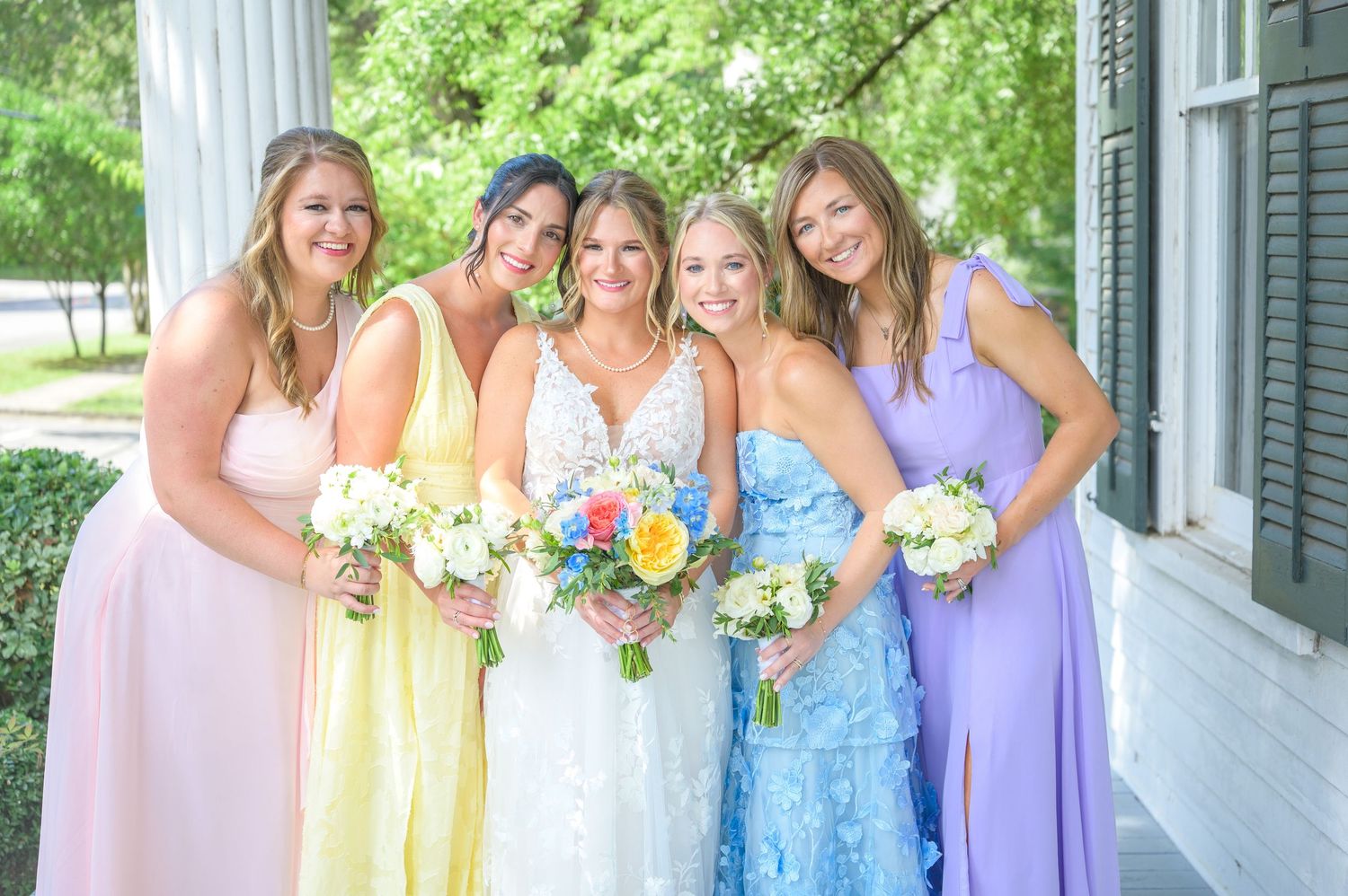Bride in white lace gown poses with four bridesmaids in pastel rainbow dresses holding bouquets outdoors.