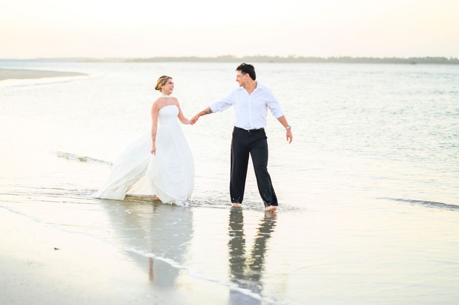 Bride and groom holding hands walking along a sandy beach shore in wedding attire at sunset.