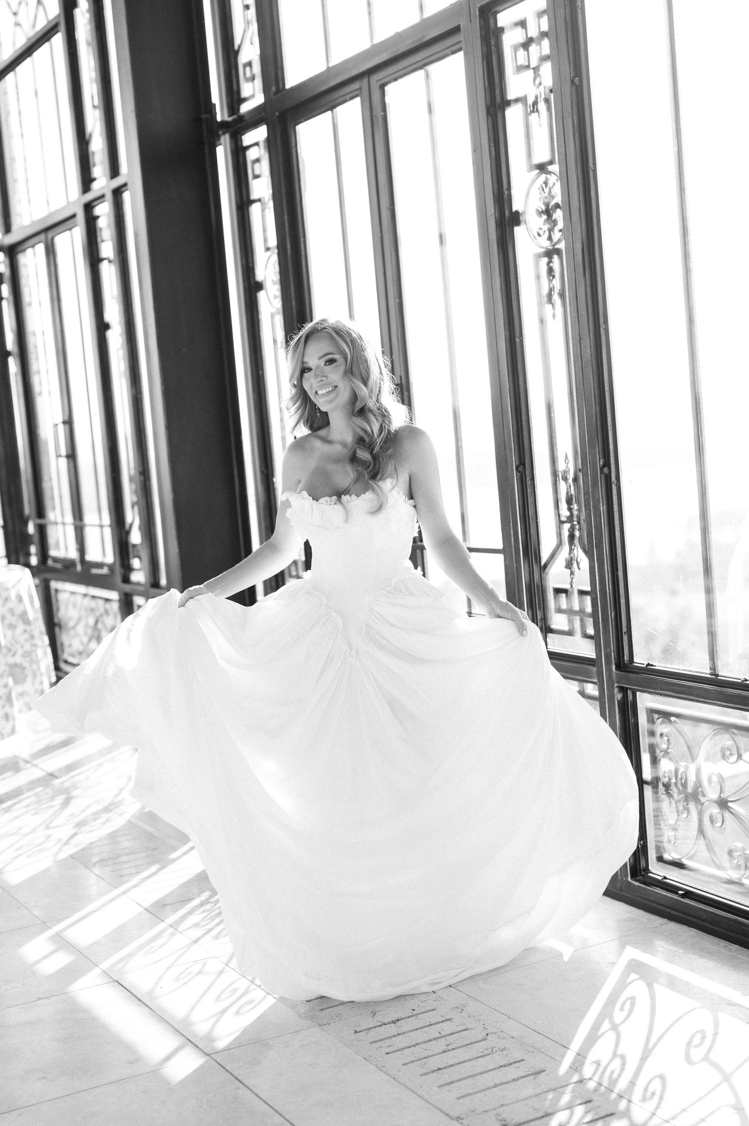 Bride twirling in a strapless ball gown wedding dress, black and white photo in an elegant venue with large windows.