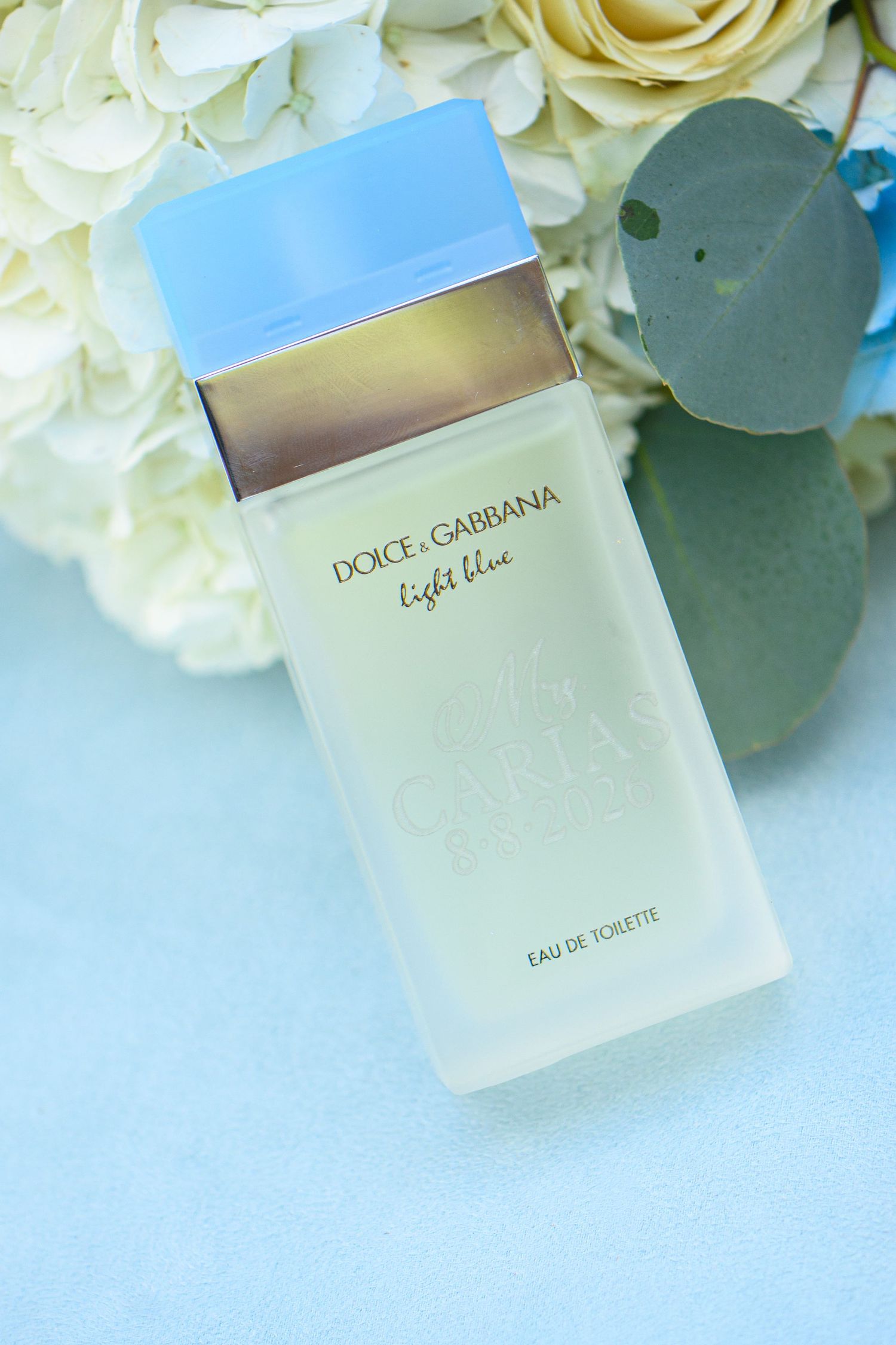 Dolce & Gabbana Light Blue Eau de Toilette bottle displayed with white roses and eucalyptus on a light blue background.