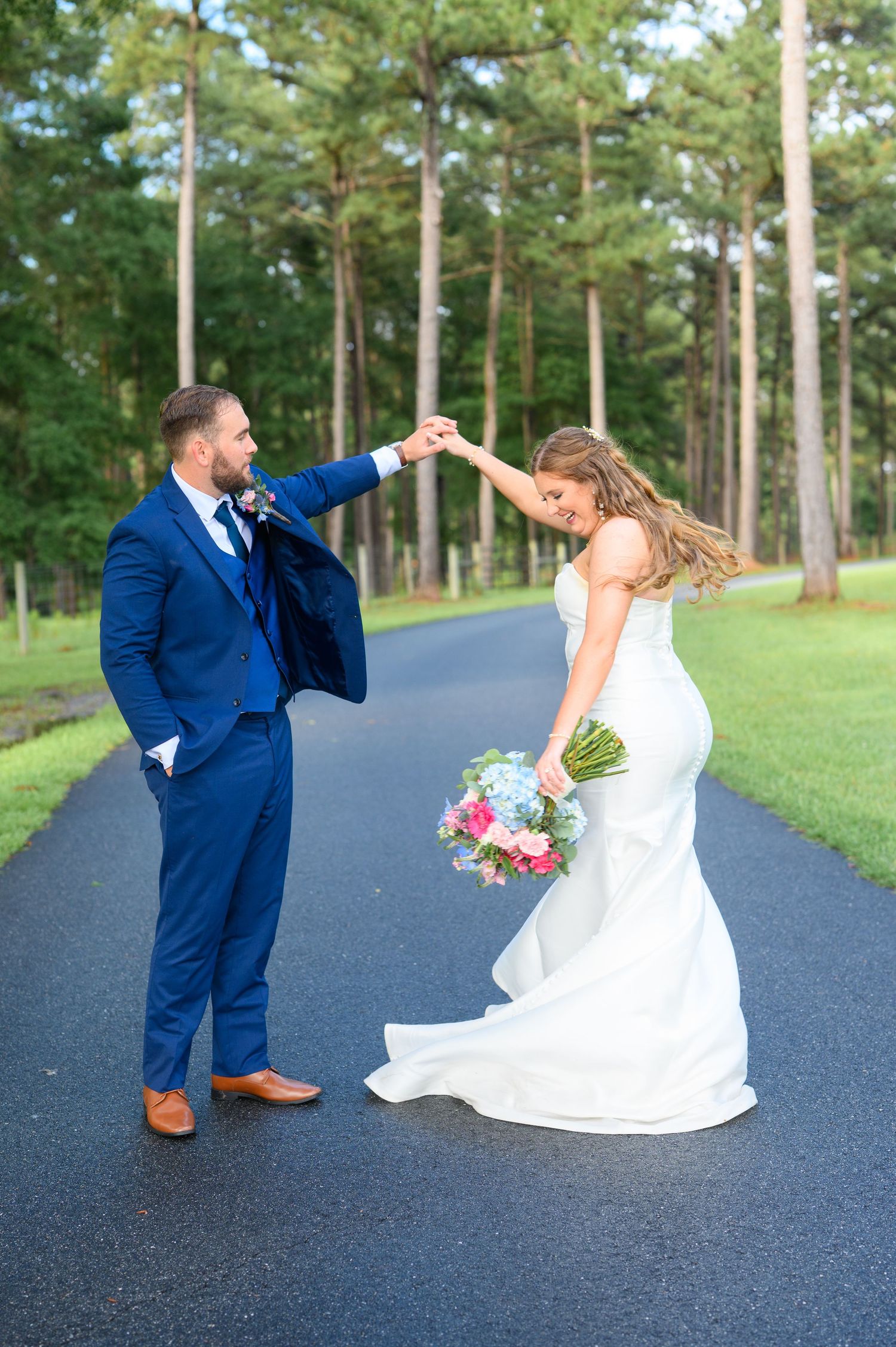 Groom in blue suit twirls bride in white mermaid gown on a tree-lined path, she holds a pink and blue bouquet.
