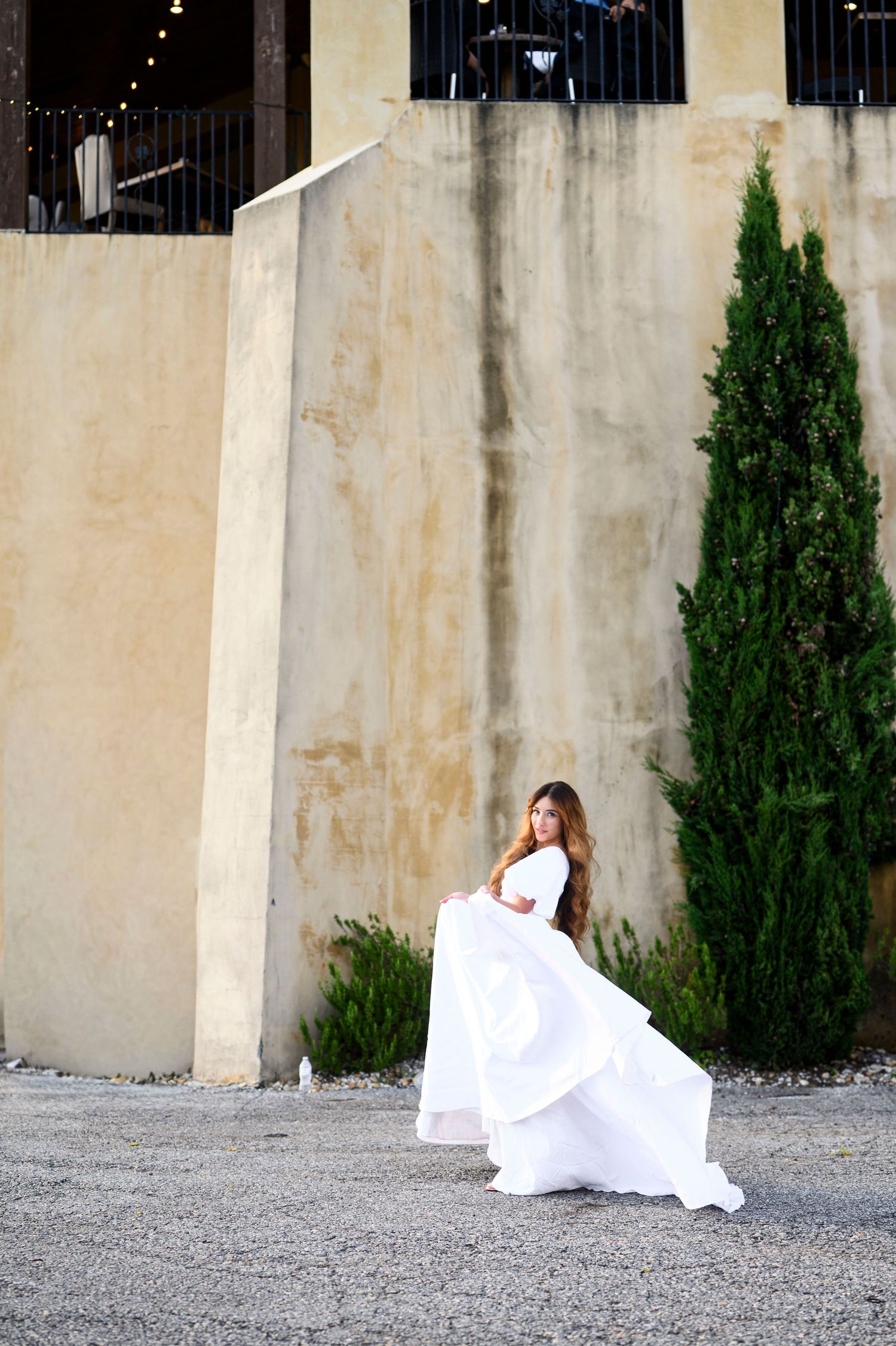 A bride in a flowing white layered gown twirls gracefully near a rustic stone wall and cypress tree outdoors.