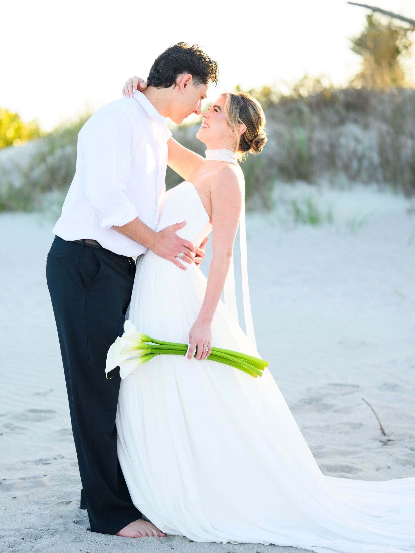 Bride and groom share a tender moment on sandy beach dunes holding calla lily bouquet.