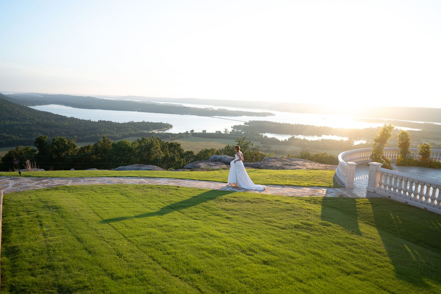 Bride in white gown stands on lush green lawn overlooking scenic lake and hills at golden hour sunset.