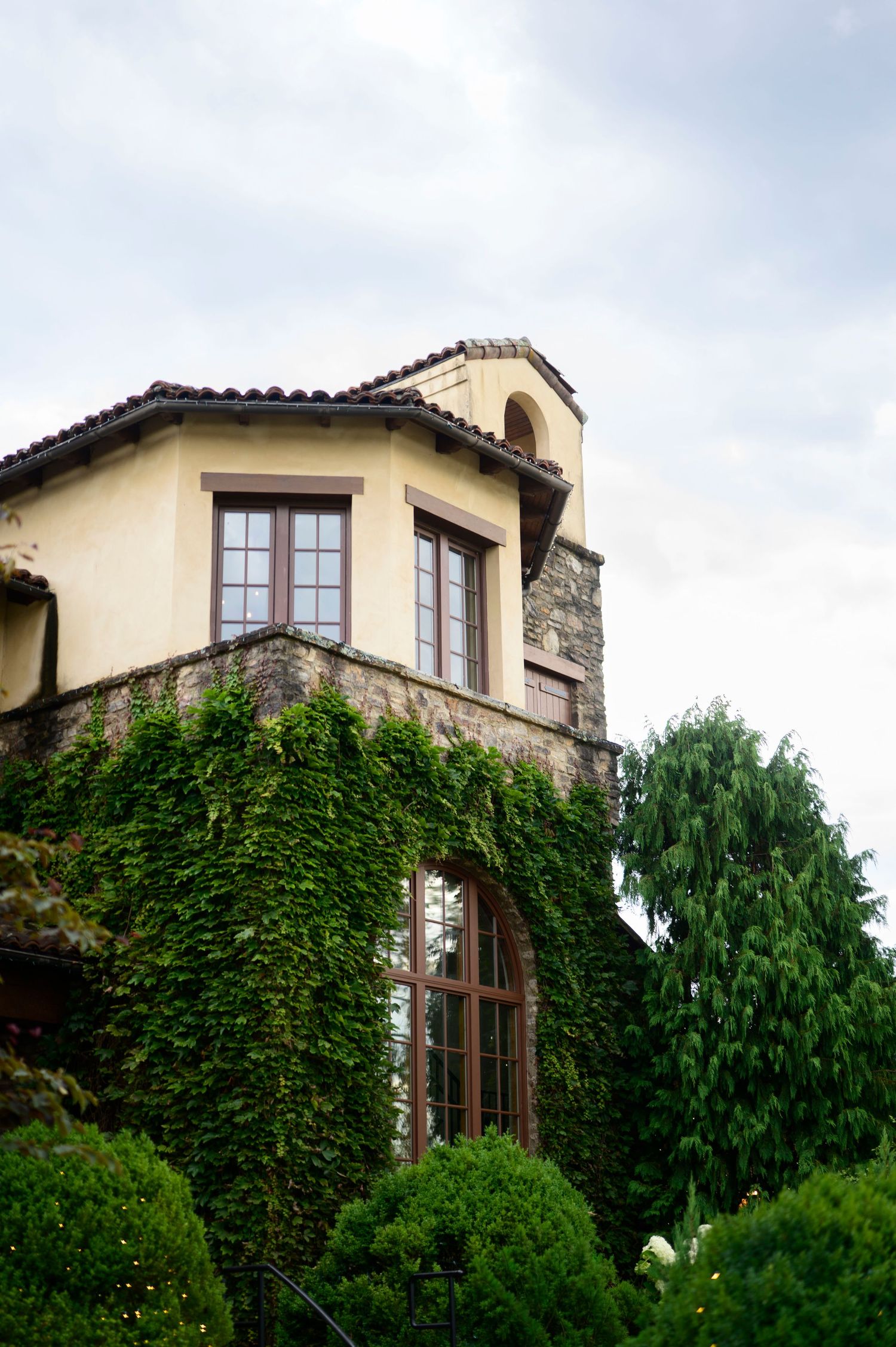 Ivy-covered stone and stucco building with arched windows, terracotta roof tiles, and lush greenery under cloudy skies.
