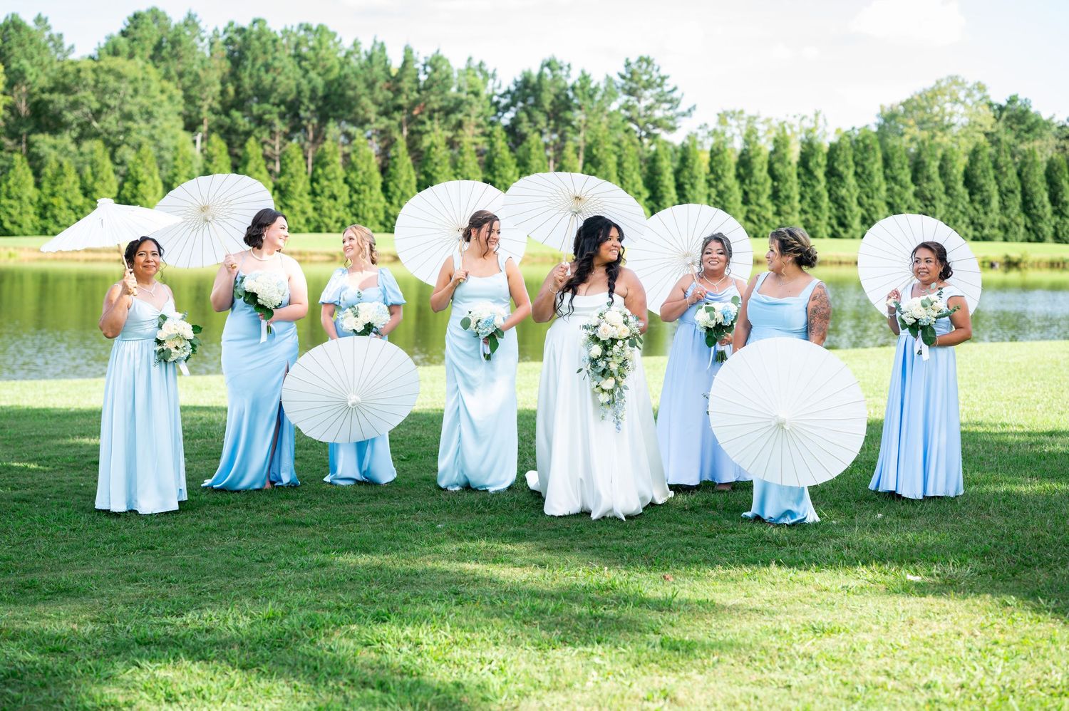 Bride and bridesmaids in light blue gowns holding white parasols on a green lawn with a pond and trees in the background.
