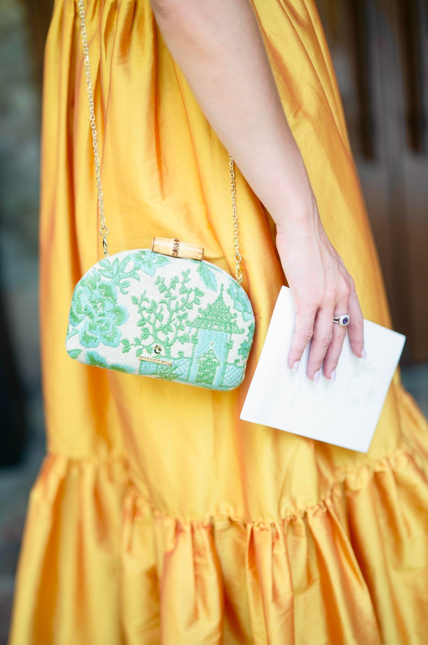 Woman in yellow dress holding white envelope beside green floral minaudière clutch bag on gold chain strap.