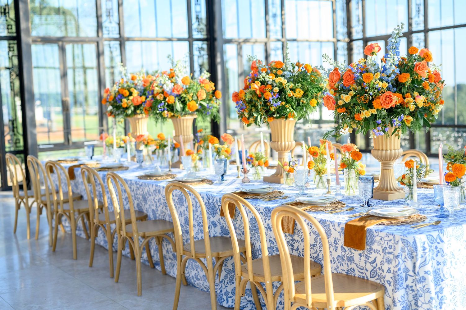 Elegant wedding reception table with blue floral linens, gold chairs, and vibrant orange floral centerpieces in a sunlit venue.