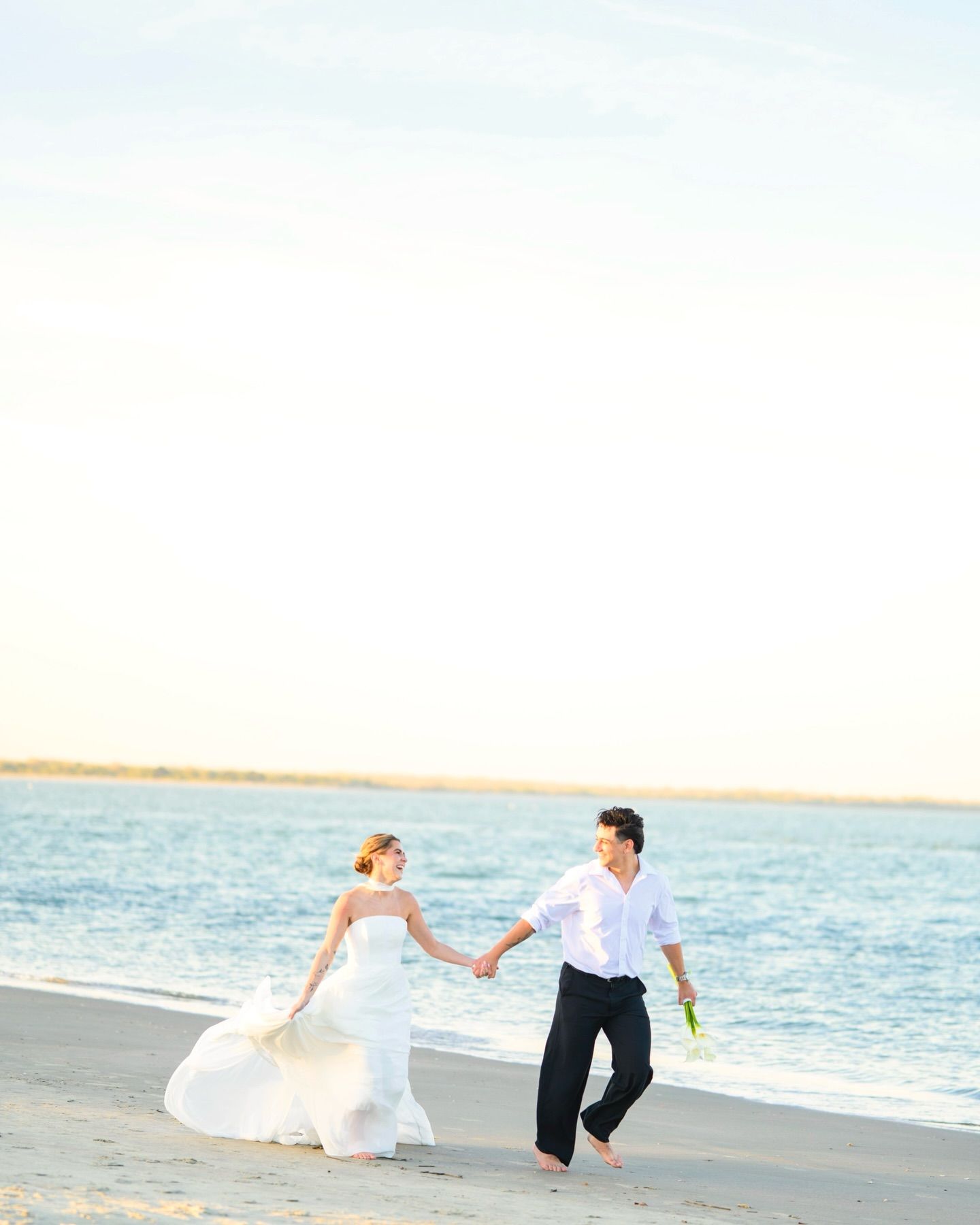 Bride and groom holding hands walking along a sunny beach at golden hour in white wedding attire.