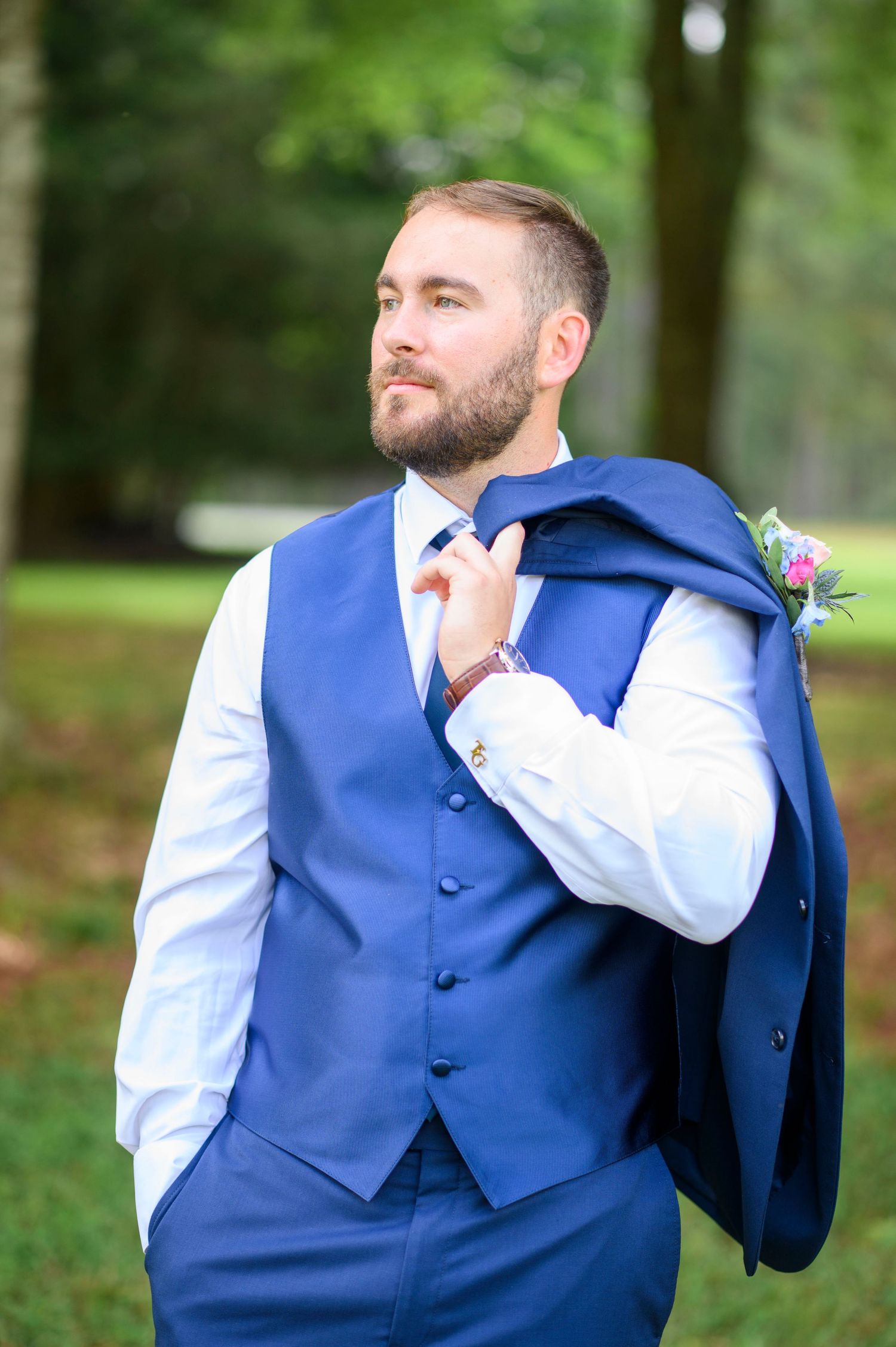 Groom in blue vest and tie holds jacket over shoulder, boutonniere visible, posing outdoors among green trees.