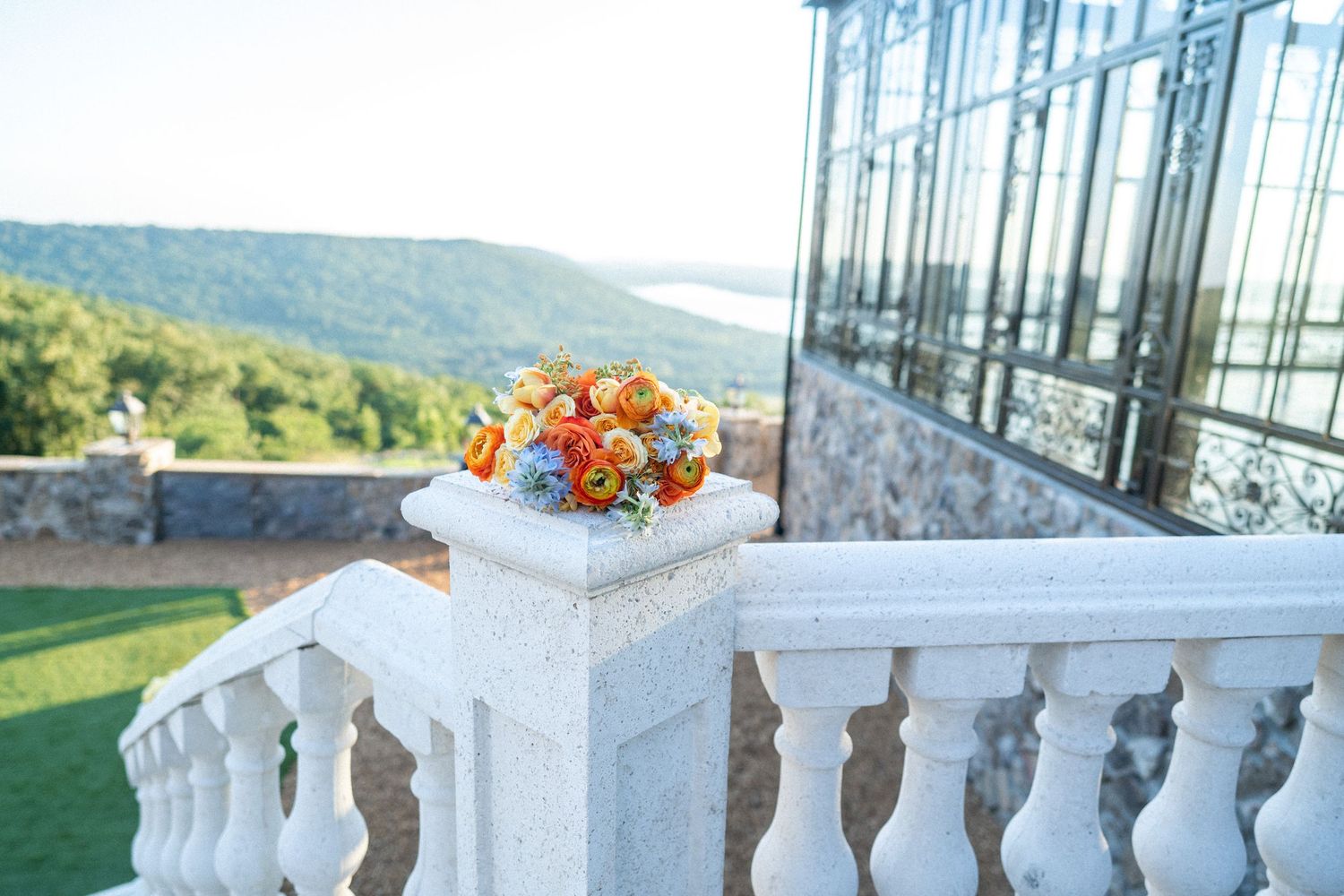 Colorful orange and blue floral bouquet on white stone balustrade with scenic Hudson Valley views and glass greenhouse.