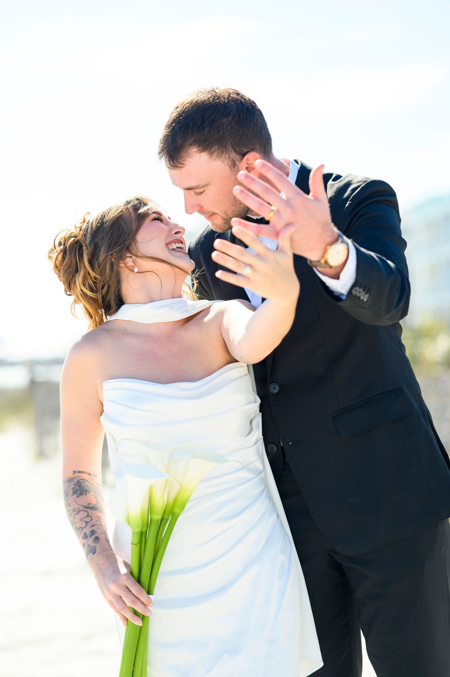 Joyful bride and groom laughing together outdoors, bride holding white calla lilies in strapless wedding dress.