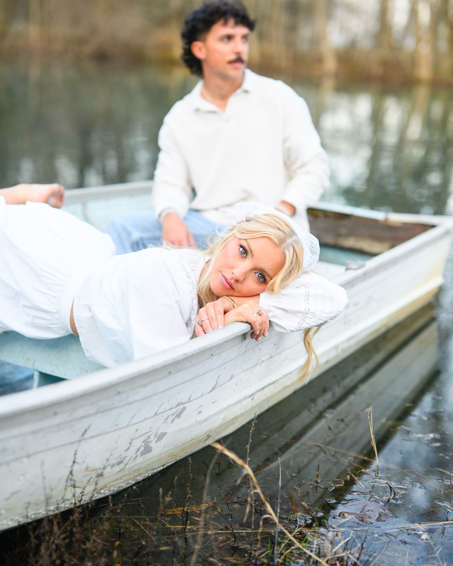 Couple in a white rowboat on a calm lake, woman lying down smiling at camera, man seated behind her.