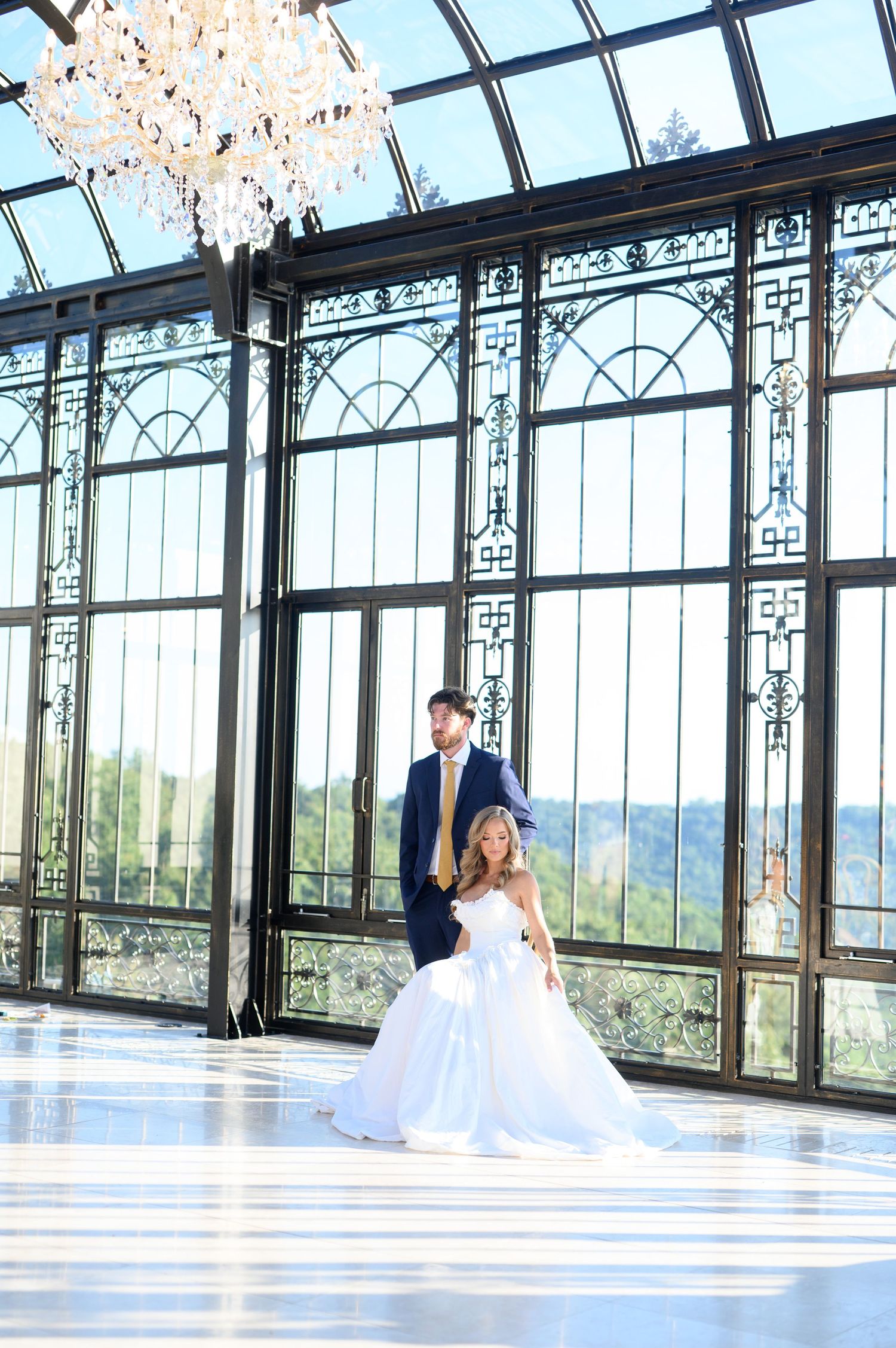 Bride and groom share a romantic moment in an elegant glass conservatory ballroom with crystal chandelier.
