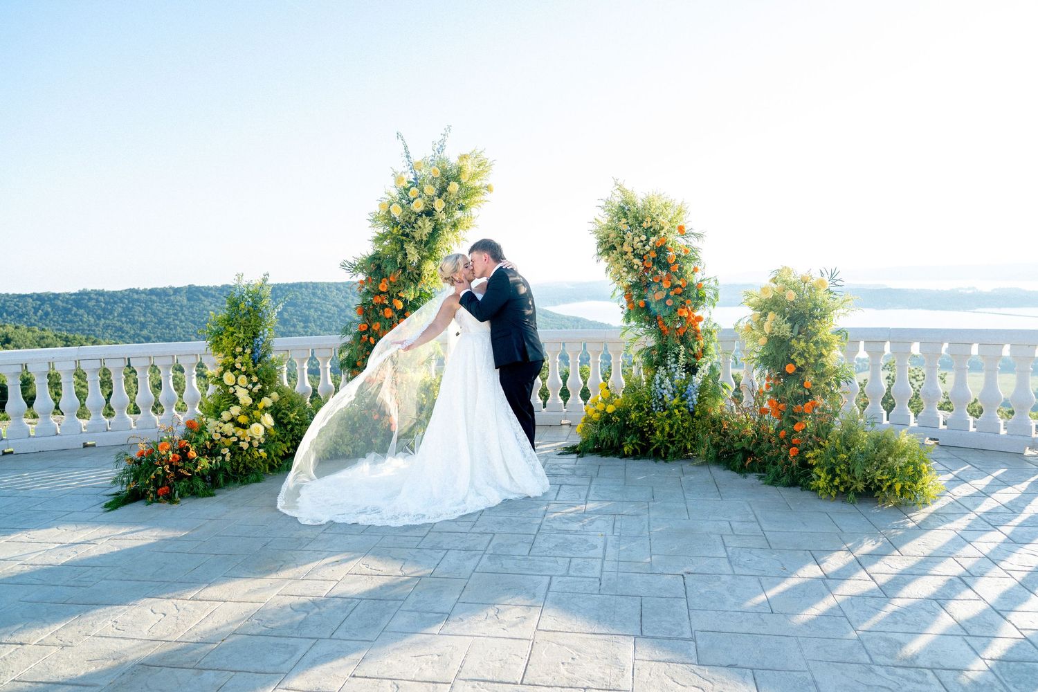 Bride and groom share a romantic kiss at an outdoor wedding ceremony with floral arch and scenic water views.