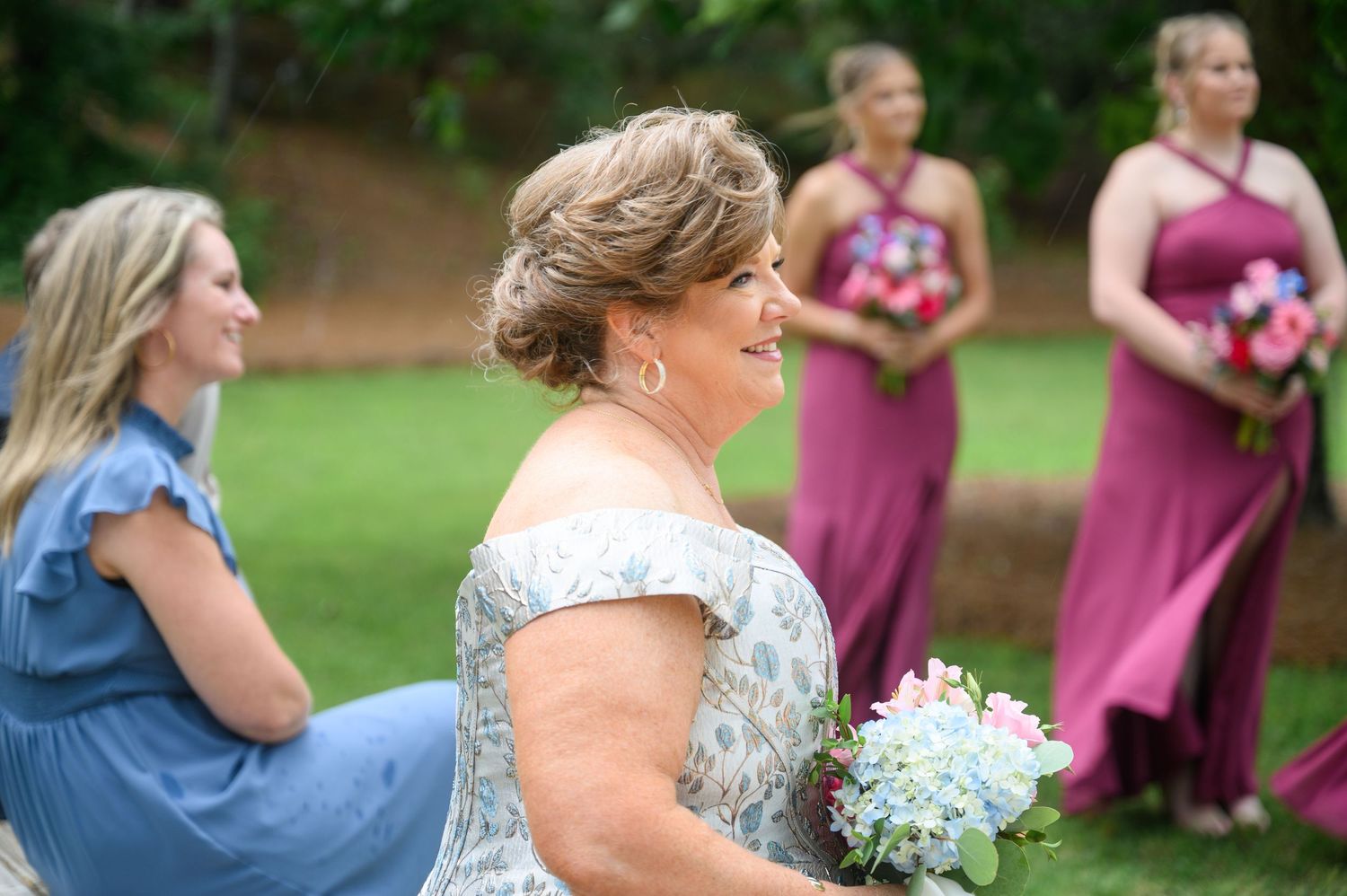 Woman in floral dress holding bouquet of blue and pink flowers at outdoor wedding with bridesmaids in mauve gowns.