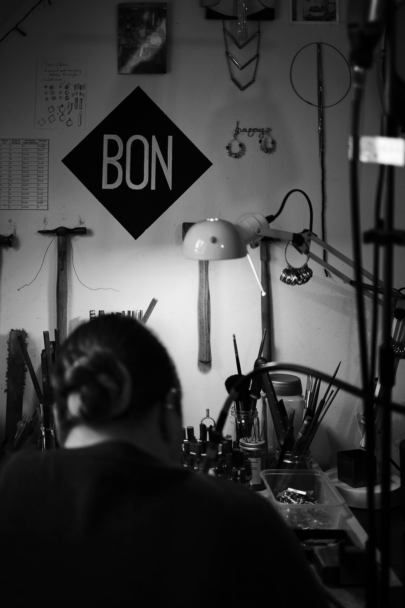 Black and white photo of a person working at a cluttered craft desk beneath a BON diamond sign and lit lamp.