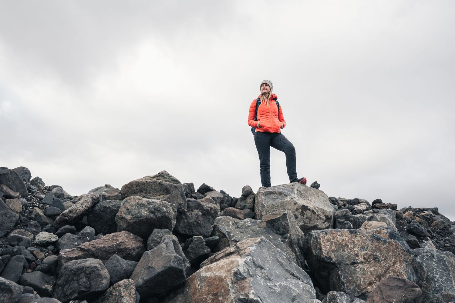 Hiker in red jacket standing triumphantly on rocky mountain summit under overcast sky.
