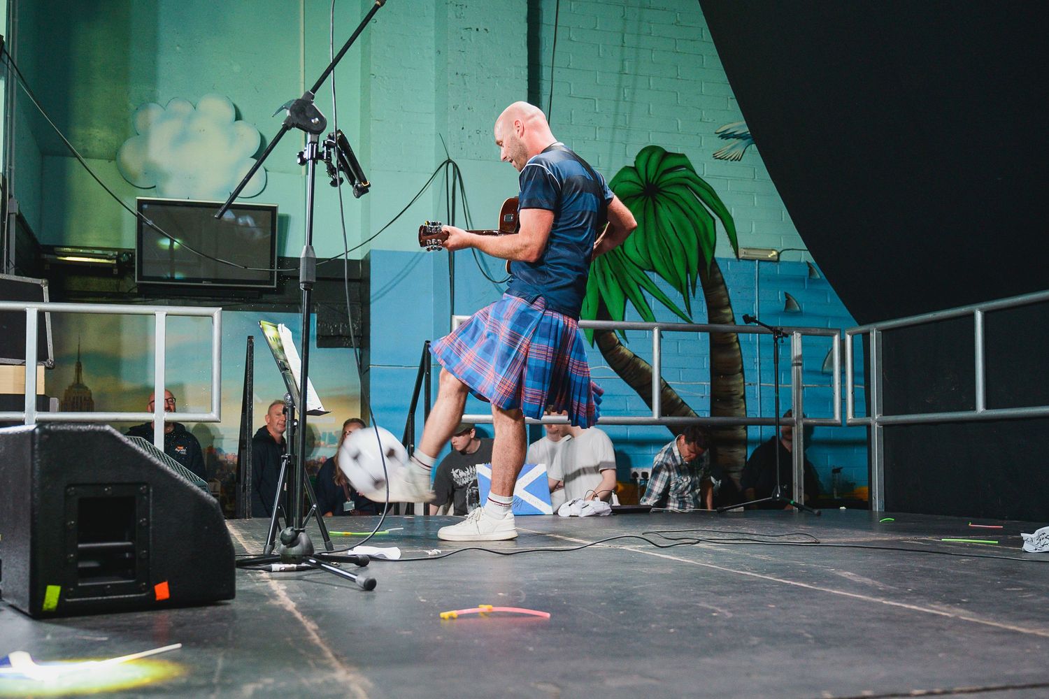 Bald man in blue shirt and tartan kilt performing on stage with speakers and palm tree backdrop.