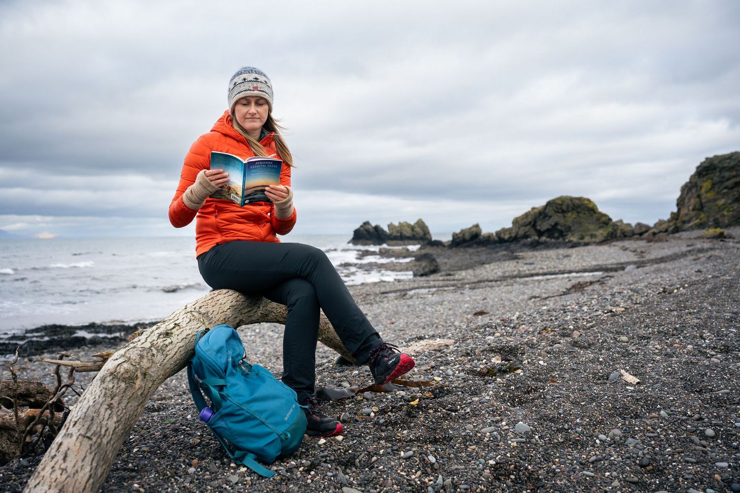 Woman in orange jacket reads book on driftwood log at rocky beach with teal backpack on overcast day.