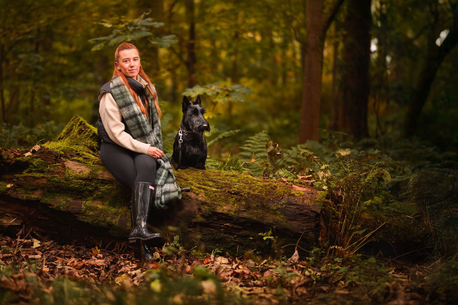 Red-haired woman sitting on a mossy log with a black dog in an autumn forest.