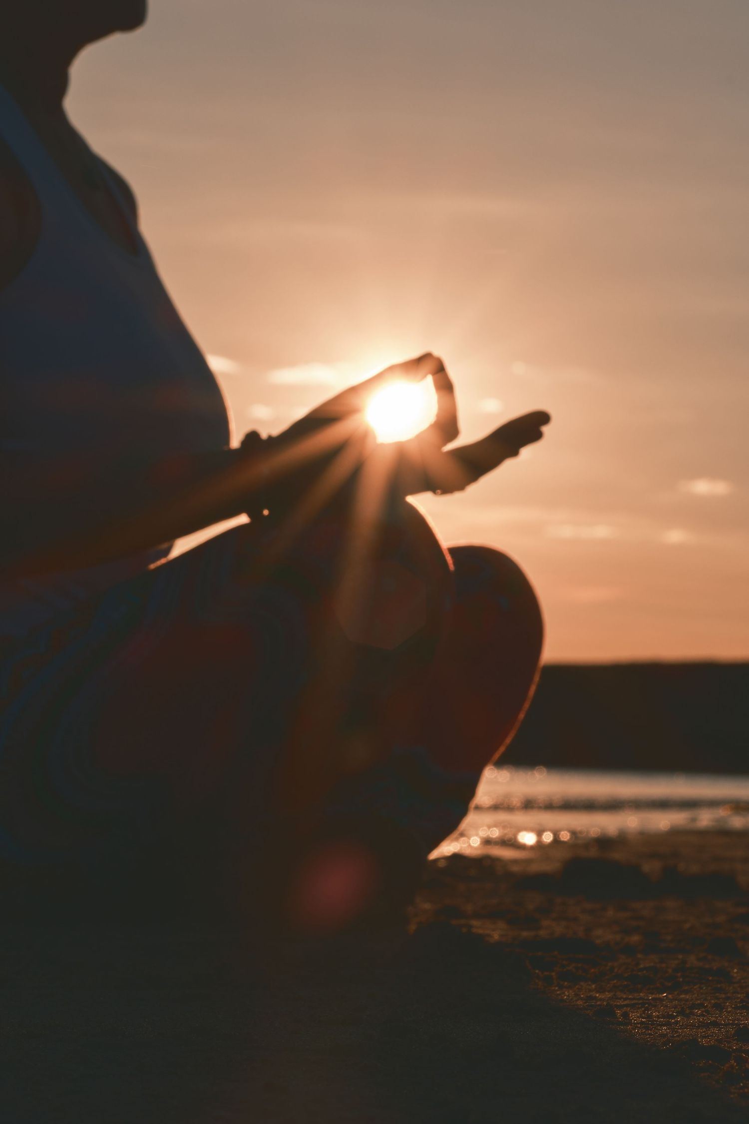 Person meditating in lotus pose on beach at sunset with sun rays glowing through fingers in serene silhouette.