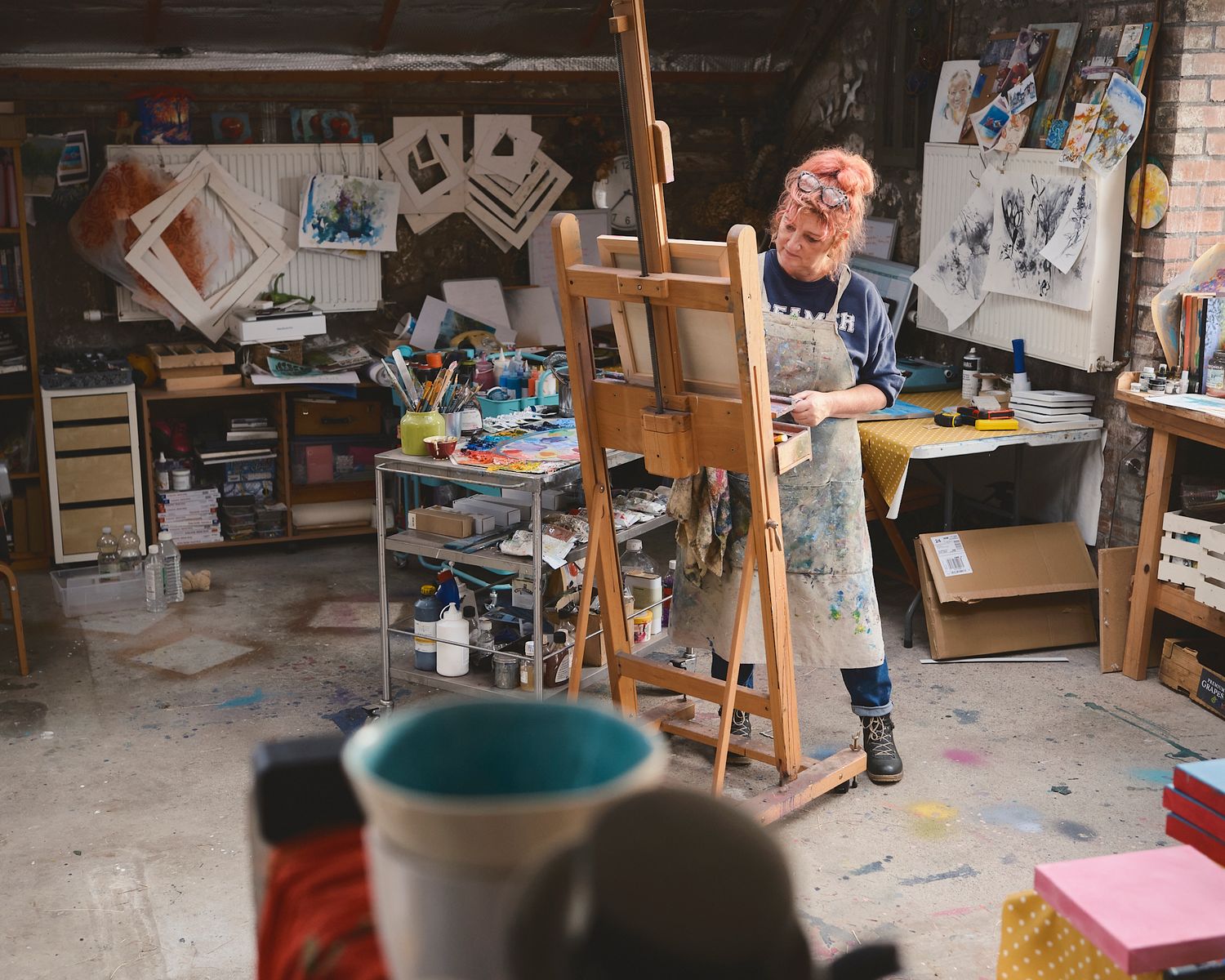 Artist working at an easel in a creative studio filled with canvases, art supplies, and colorful paint splatters.