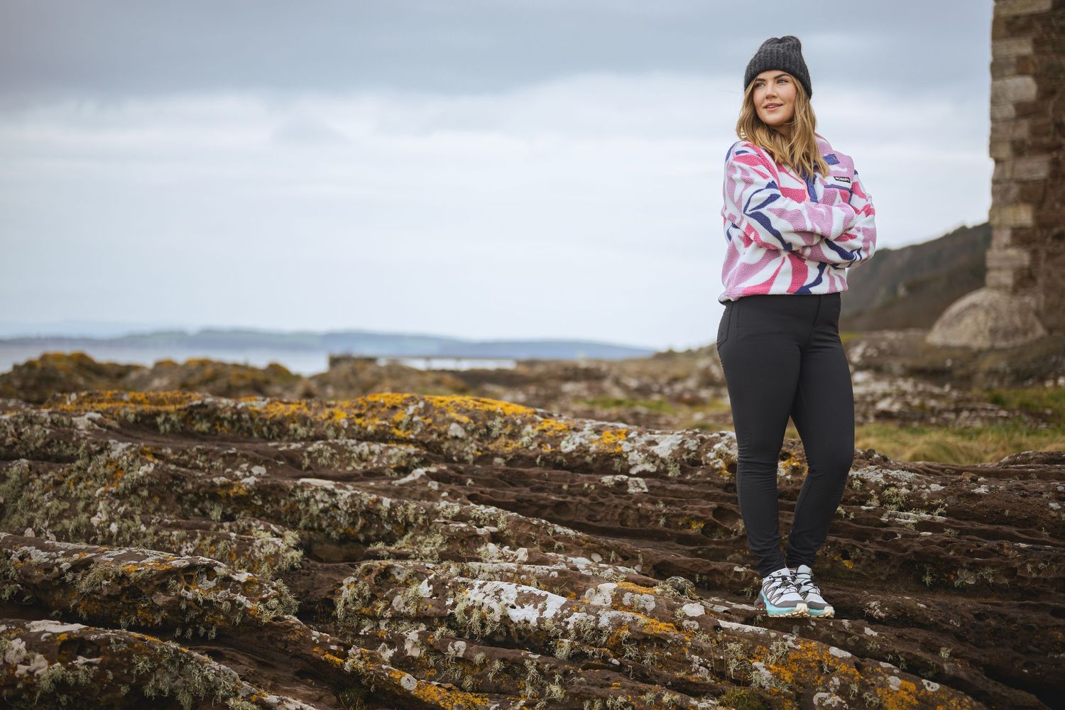 Woman in pink hoodie and black pants stands on rocky coastal shore with arms crossed on overcast day.