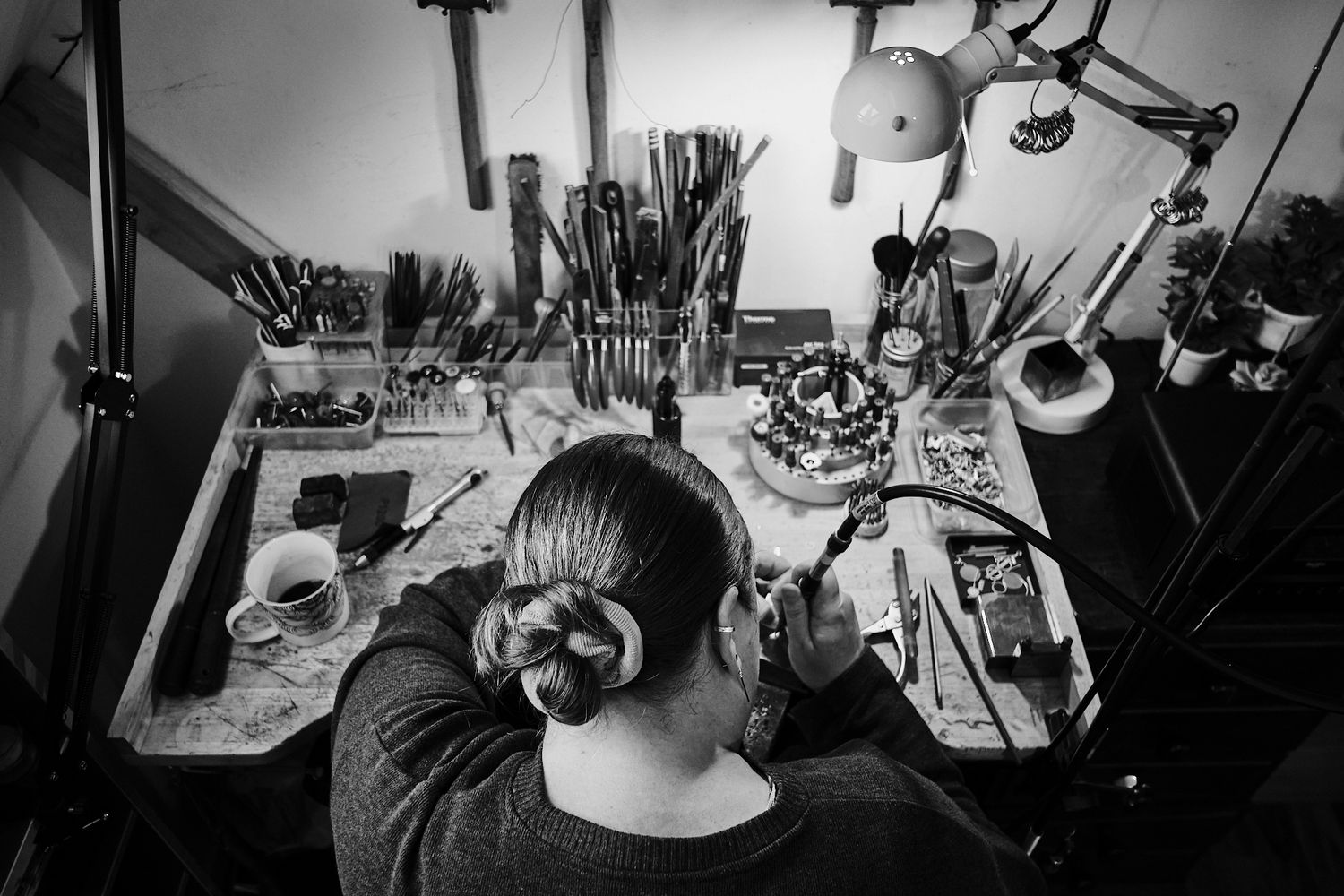 Black and white overhead view of a female jeweler working at a cluttered workbench with tools.