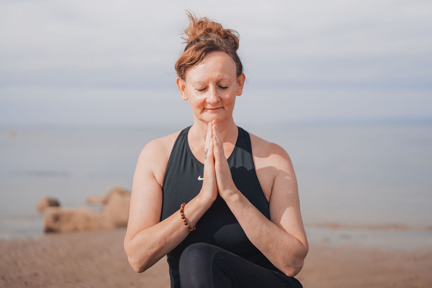 Woman in black yoga attire meditating with hands in prayer pose on a serene beach setting.