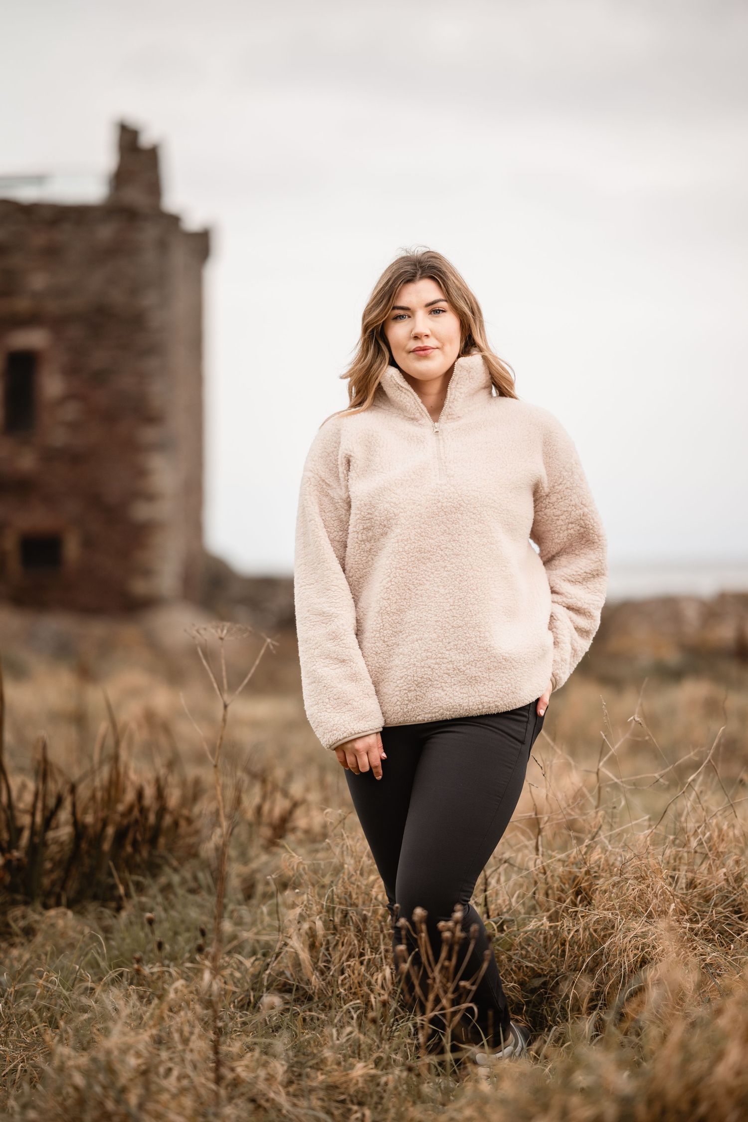 Woman in cream fleece pullover and black leggings standing in a field with a ruined stone tower behind her.