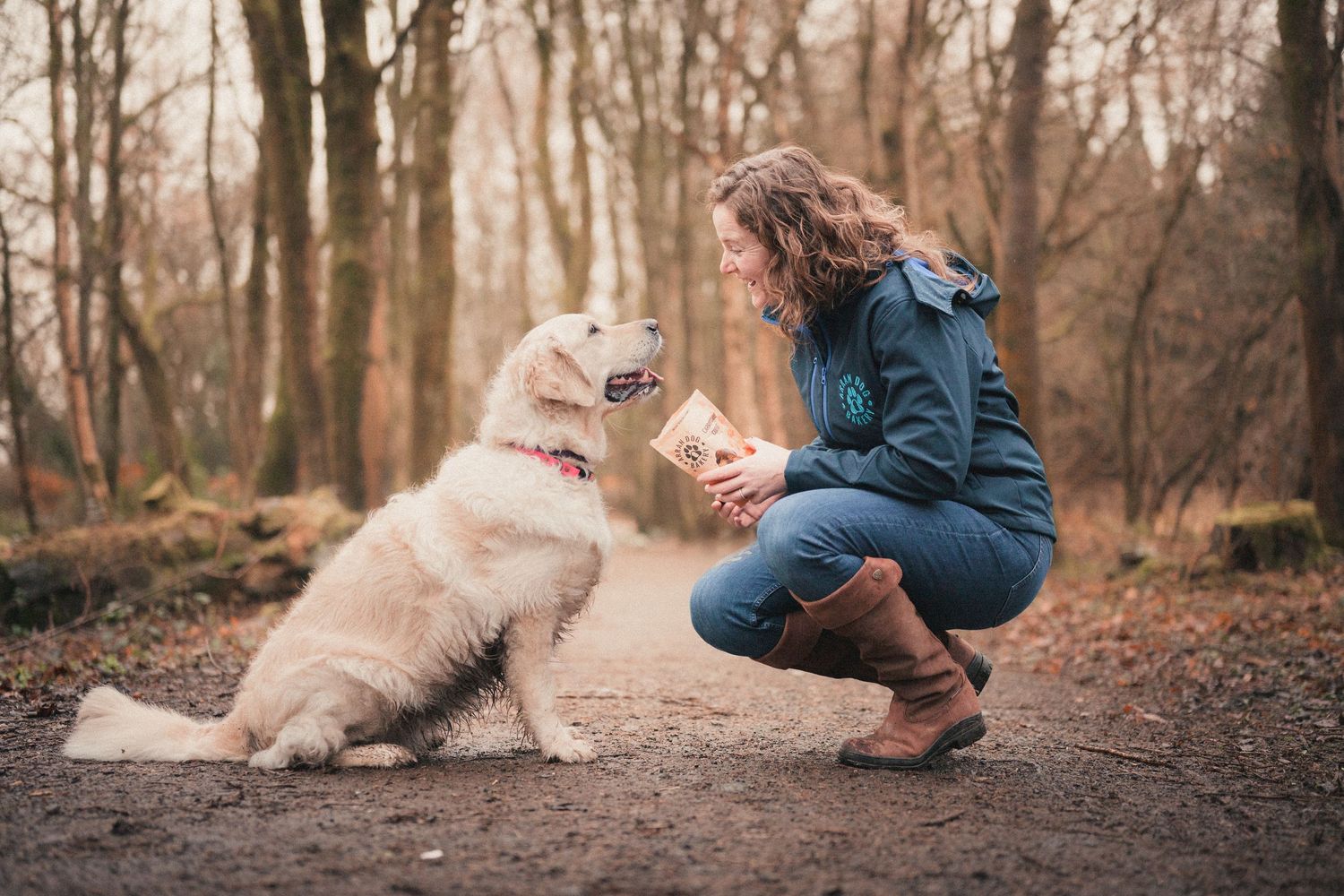 Woman crouching on forest path feeding treat to fluffy white dog in autumn woodland setting.