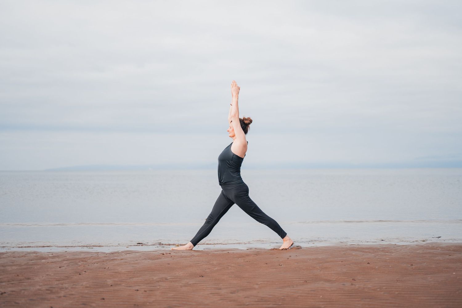 Woman practicing yoga warrior pose on a sandy beach with calm ocean and overcast sky in the background.